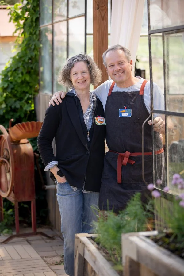 A smiling woman and man with a nickname badge that says 'Eric' in a greenhouse surrounded by plants, with the woman wearing a jacket and the man wearing an apron.