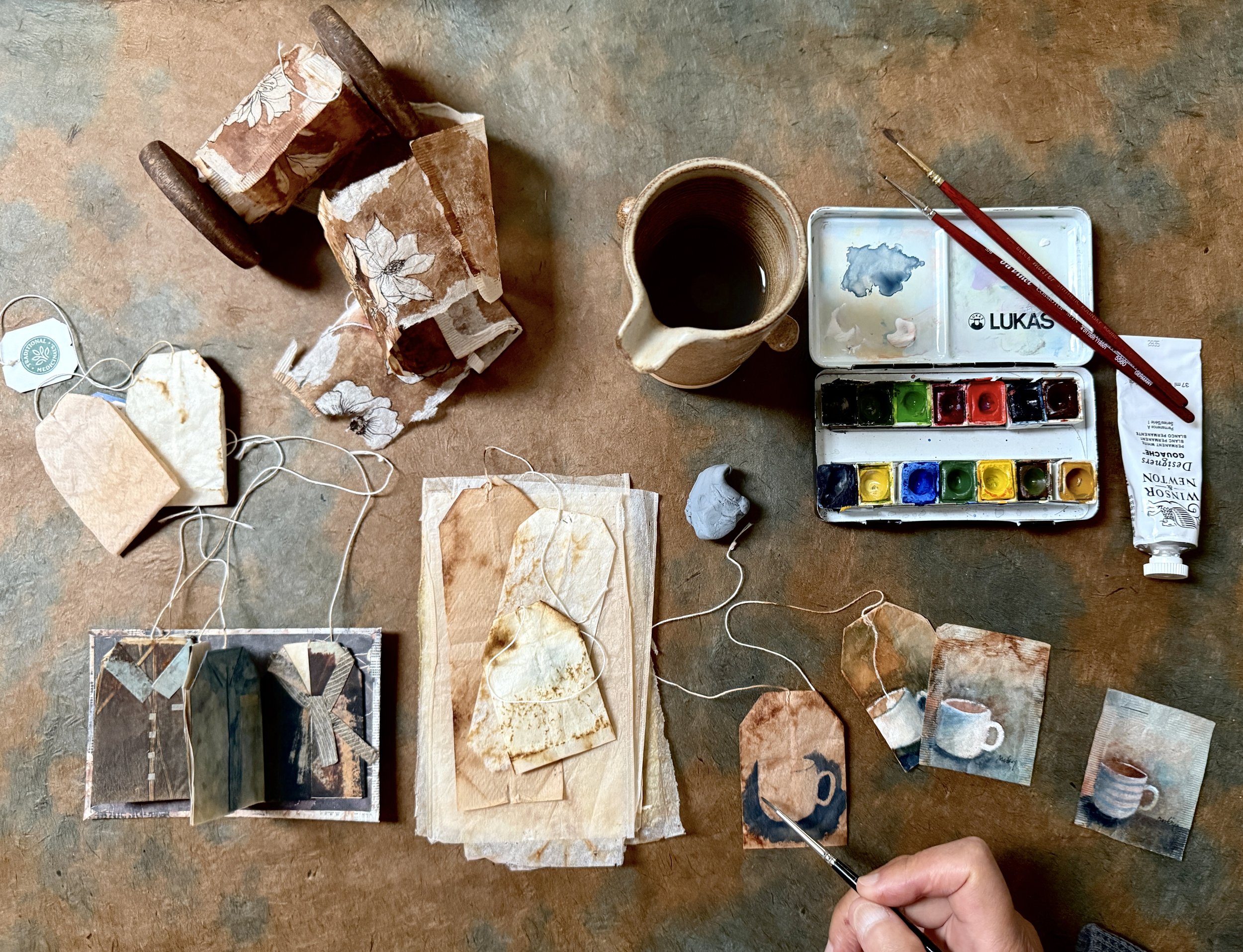 Art supplies and small fabric samples on a wooden surface, including a watercolor palette, brush, ceramic mug of water, and pieces of fabric with various patterns and textures.
