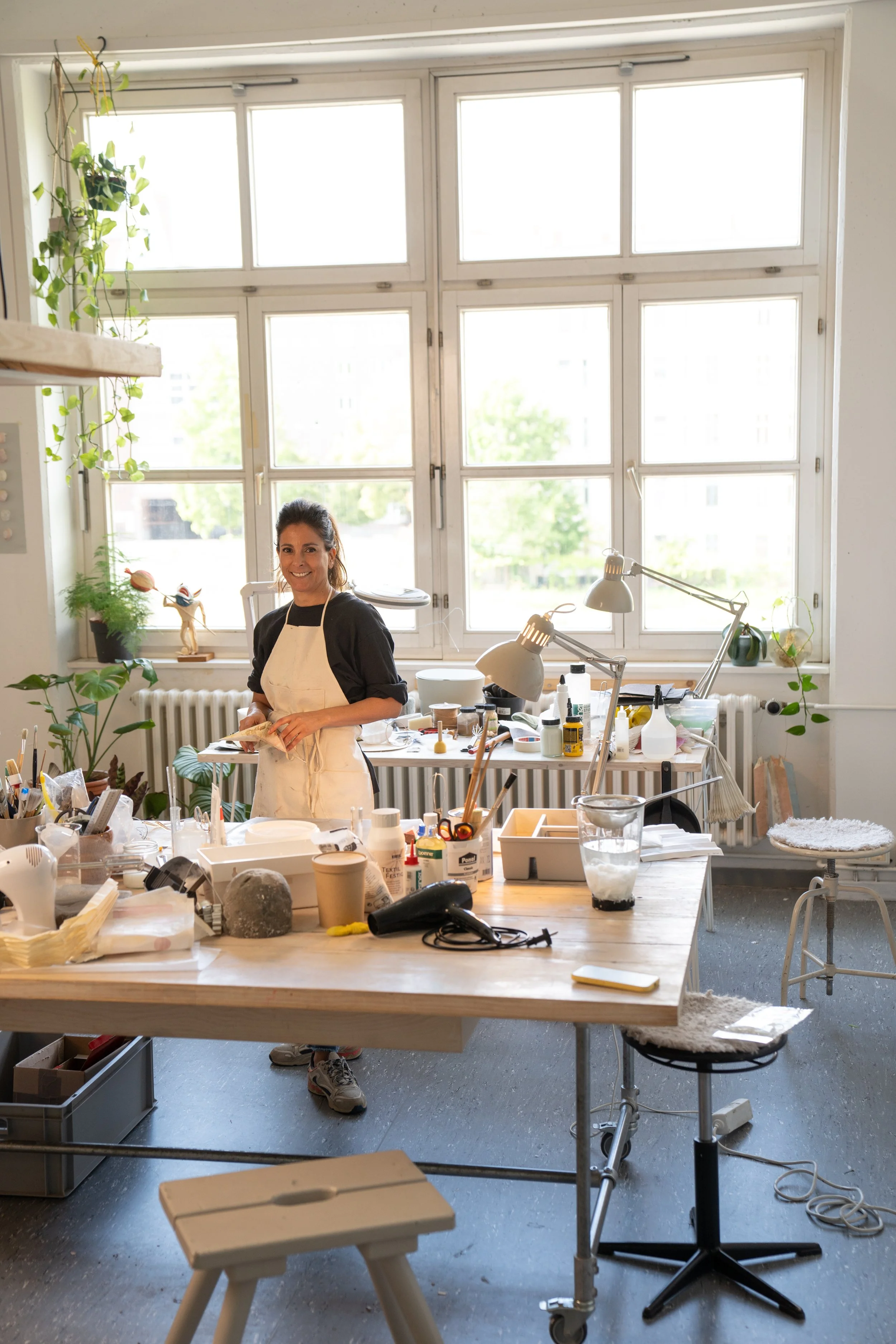 A woman in a black shirt and white apron smiling in an art studio filled with art supplies, plants, and bright large windows.