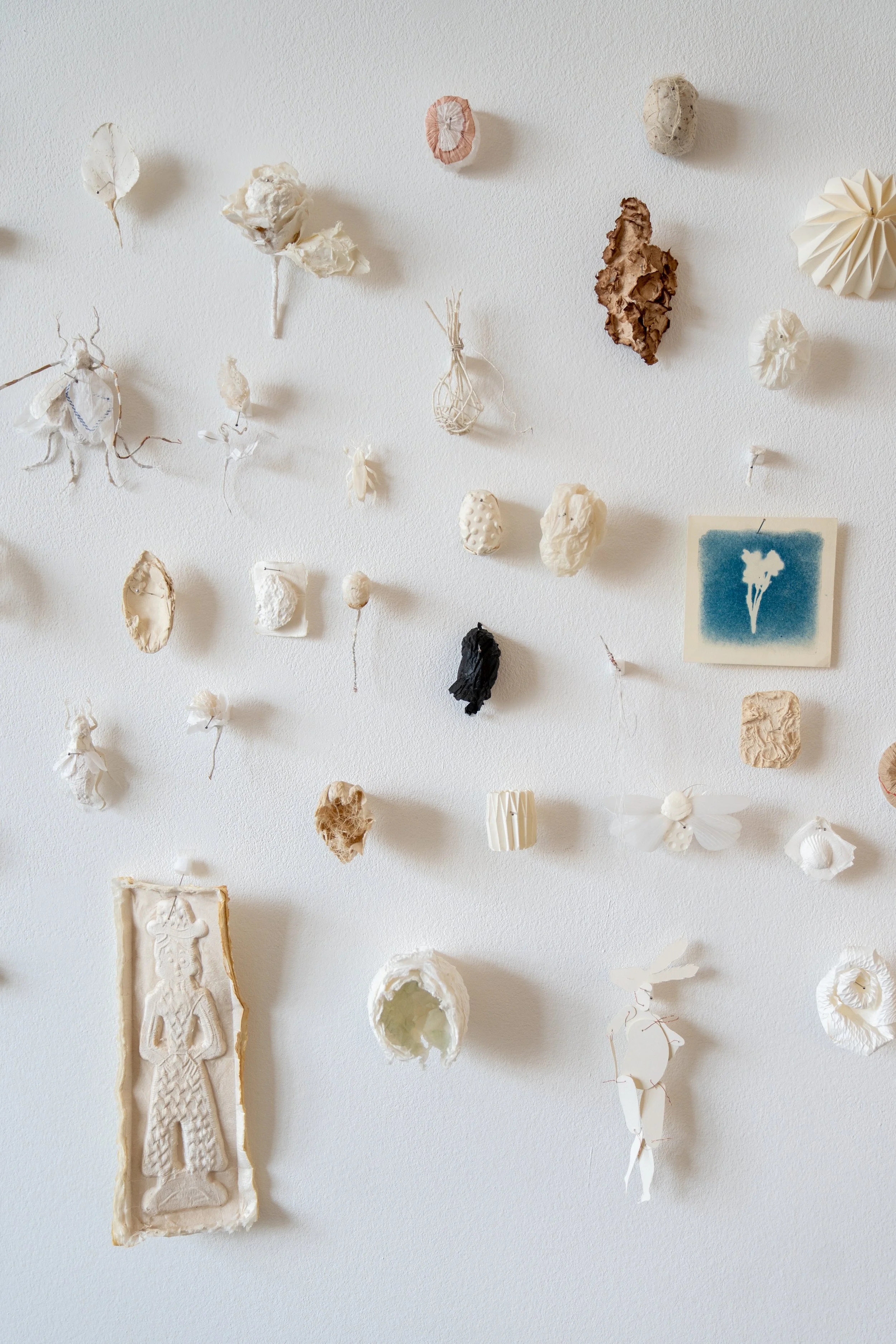 Wall decorated with various small paper and textile art pieces, including a blue ink print of a flower, a carved figure, and textured shapes.