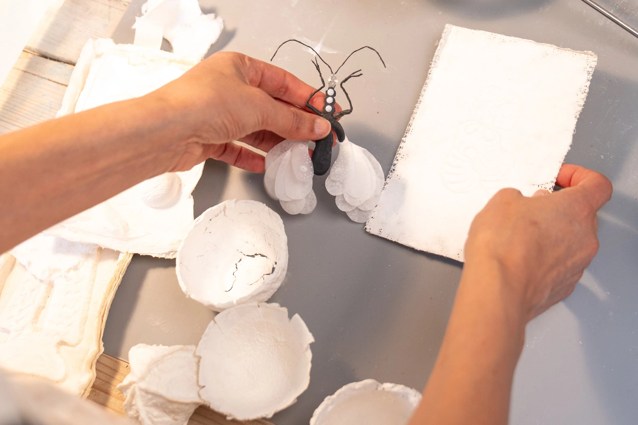 A person is holding a paper cutout butterfly with black outlines and white dots on a metallic surface. Around the hand, there are several white paper or plaster pieces and torn sheets.