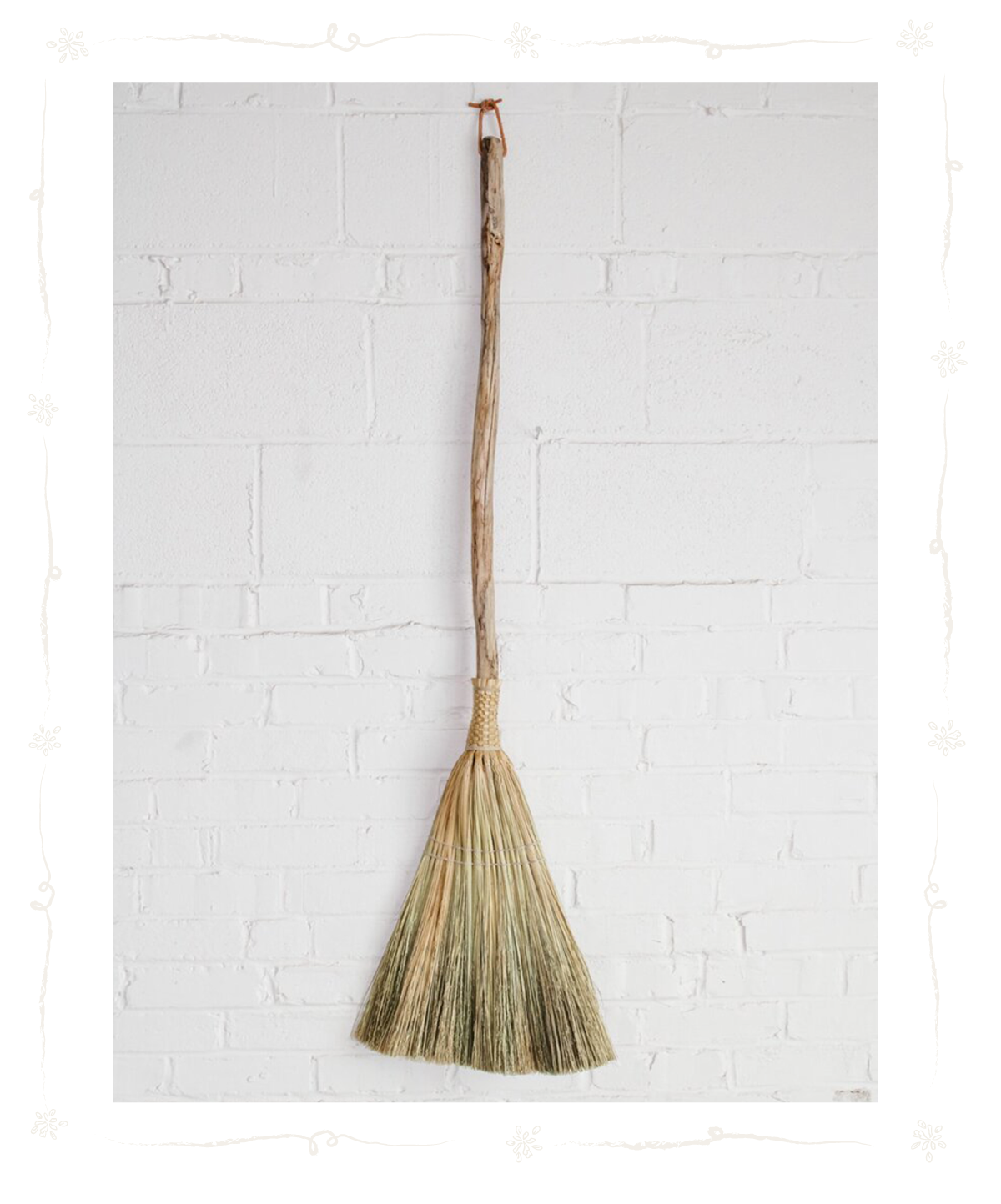 A traditional straw broom with a wooden handle hanging on a white brick wall.
