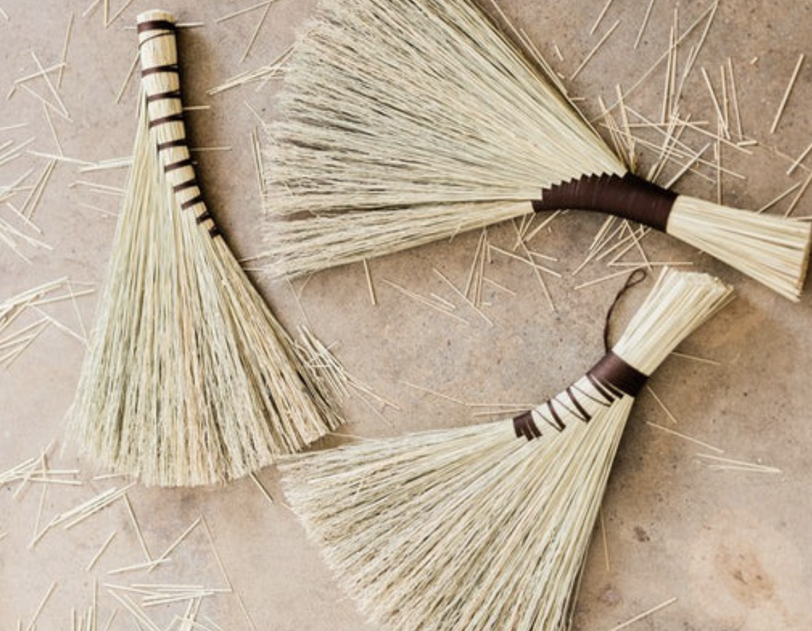Two small handmade brooms with natural bristles, wrapped with brown string, are on a tiled surface surrounded by scattered straw or wood sticks.