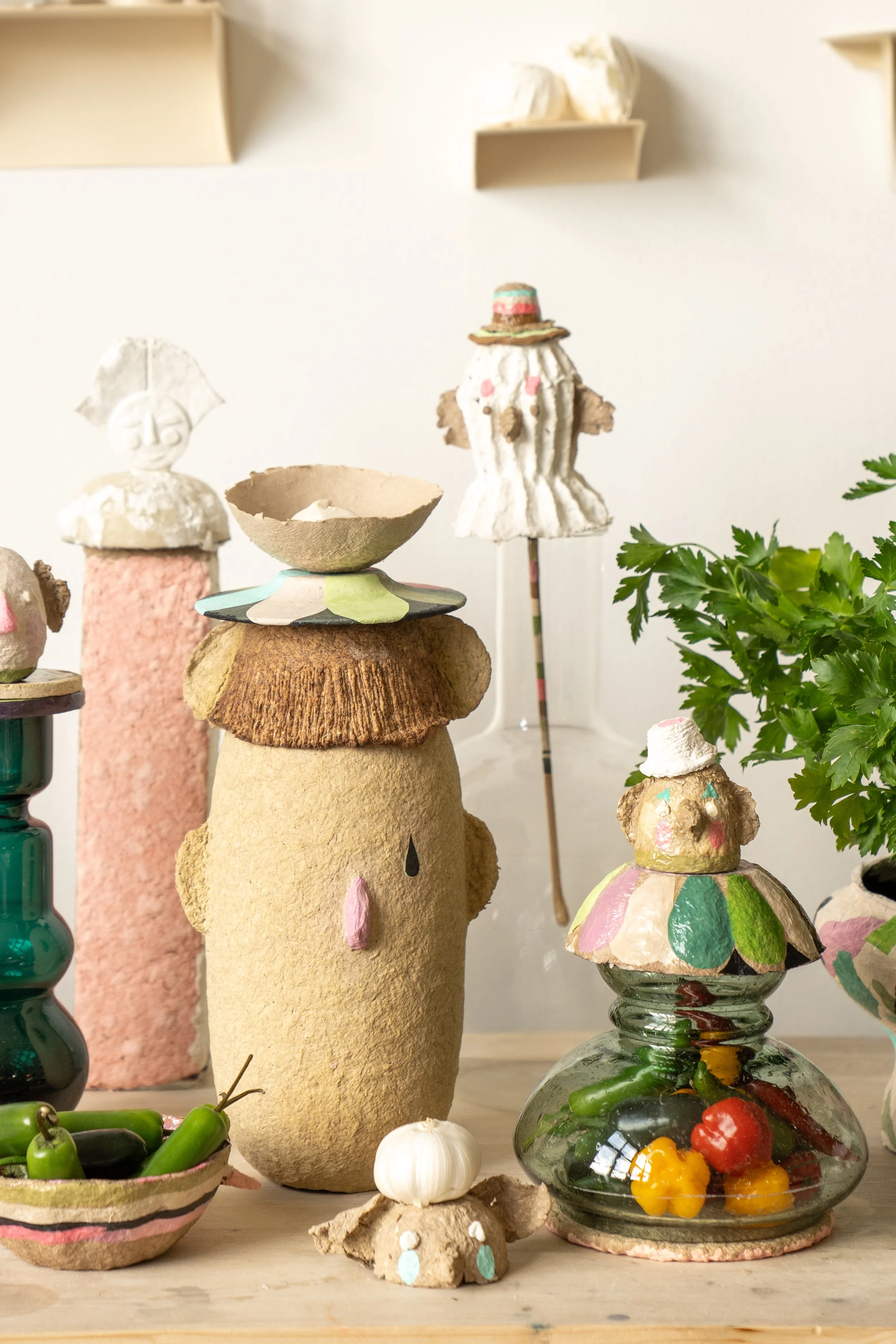 Various handcrafted figurines and pottery, including a face with a pink nose, a bowl, and decorative objects crafted with clay and paint, with a greenery plant on the right side.