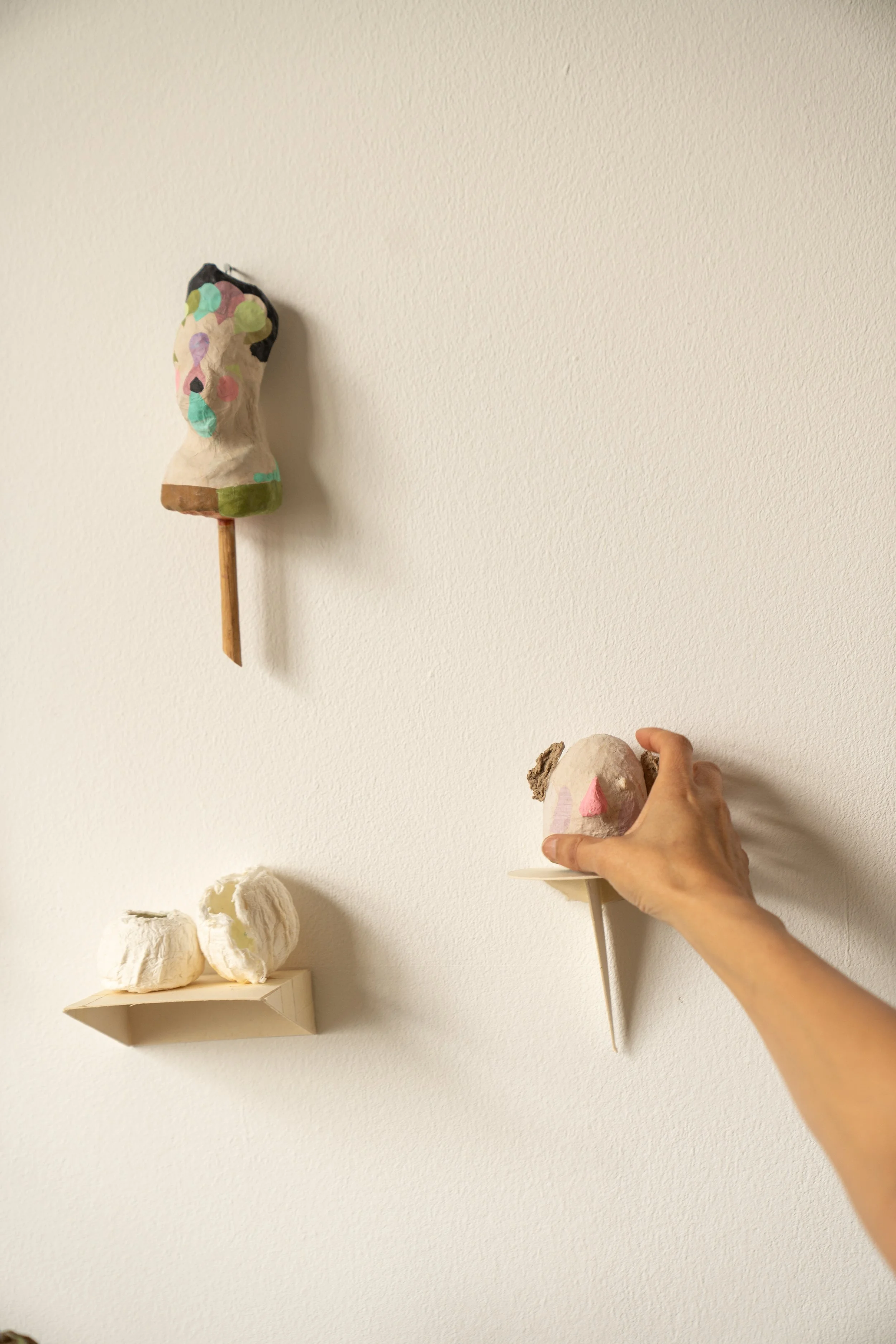 Three soft sculptures resembling faces placed on shelves and a wall, with a hand adjusting one sculpture in the lower right corner of the image.