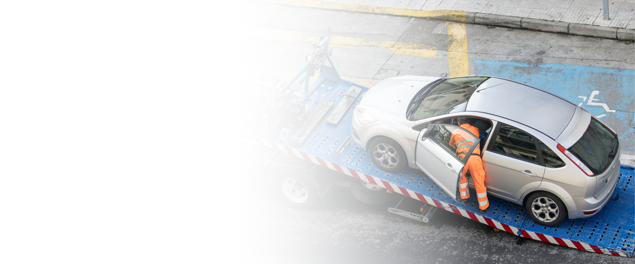 Workers loading a silver car onto a flatbed tow truck on an urban street with parking lines and a blue accessible parking spot.