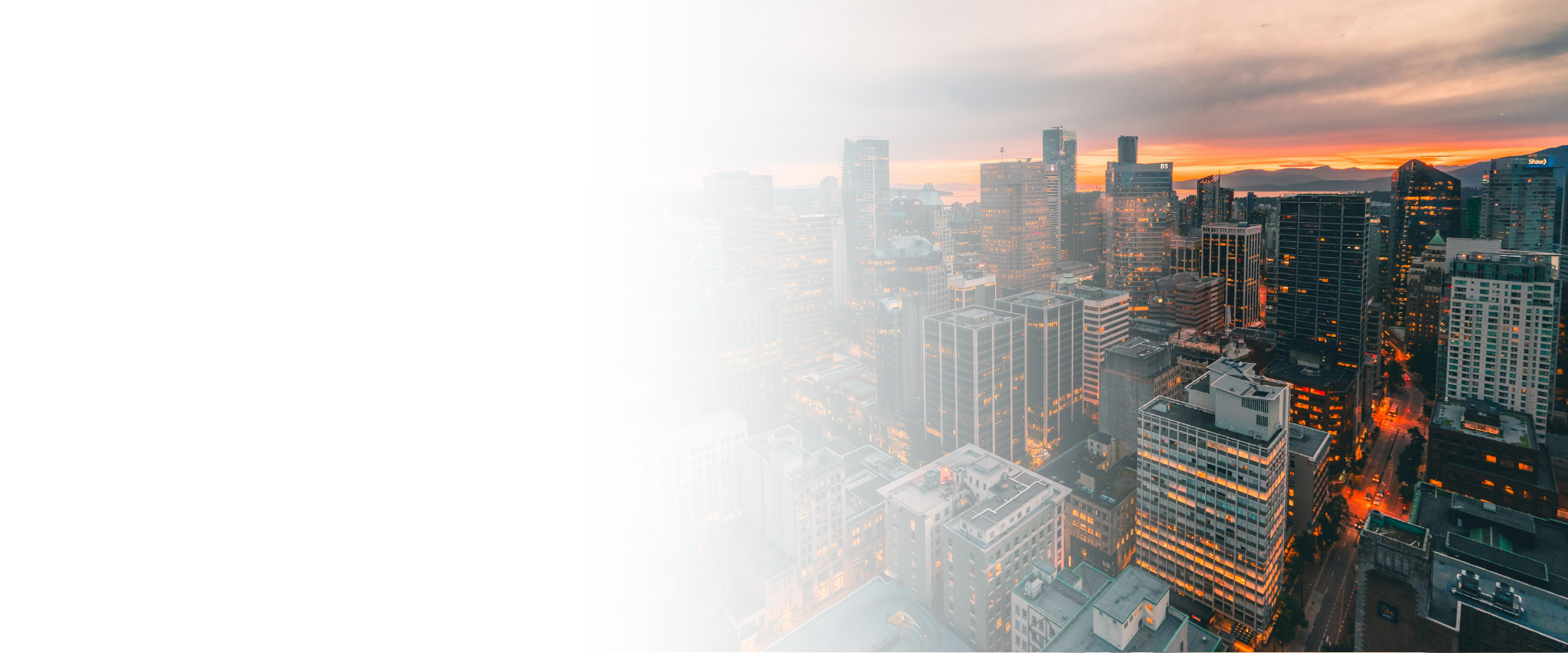 A city skyline at sunset with tall buildings, some with lit windows, and mountains in the background.