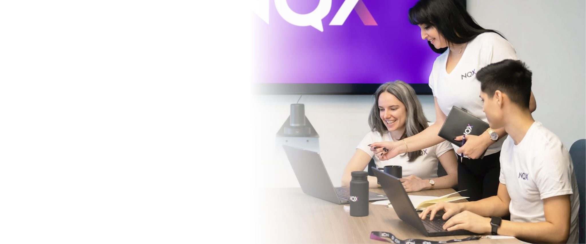 Three women and one man working together in a meeting room, with laptops, notebooks, and water bottles on the table, and a large screen displaying the ''NQX'' logo behind them.