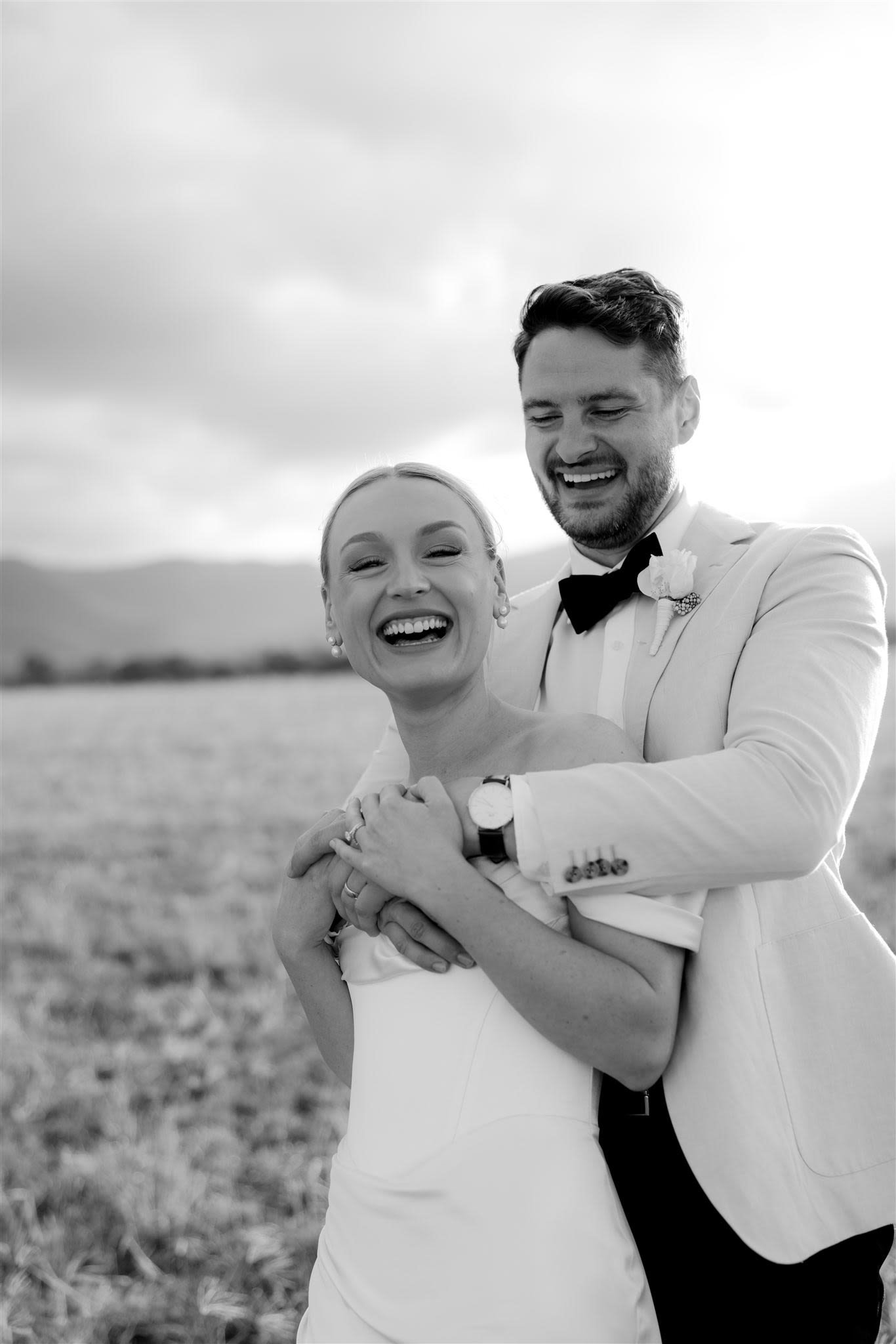 Black and white photo of a joyful couple in formal attire, with a man embracing a woman from behind in an open field.