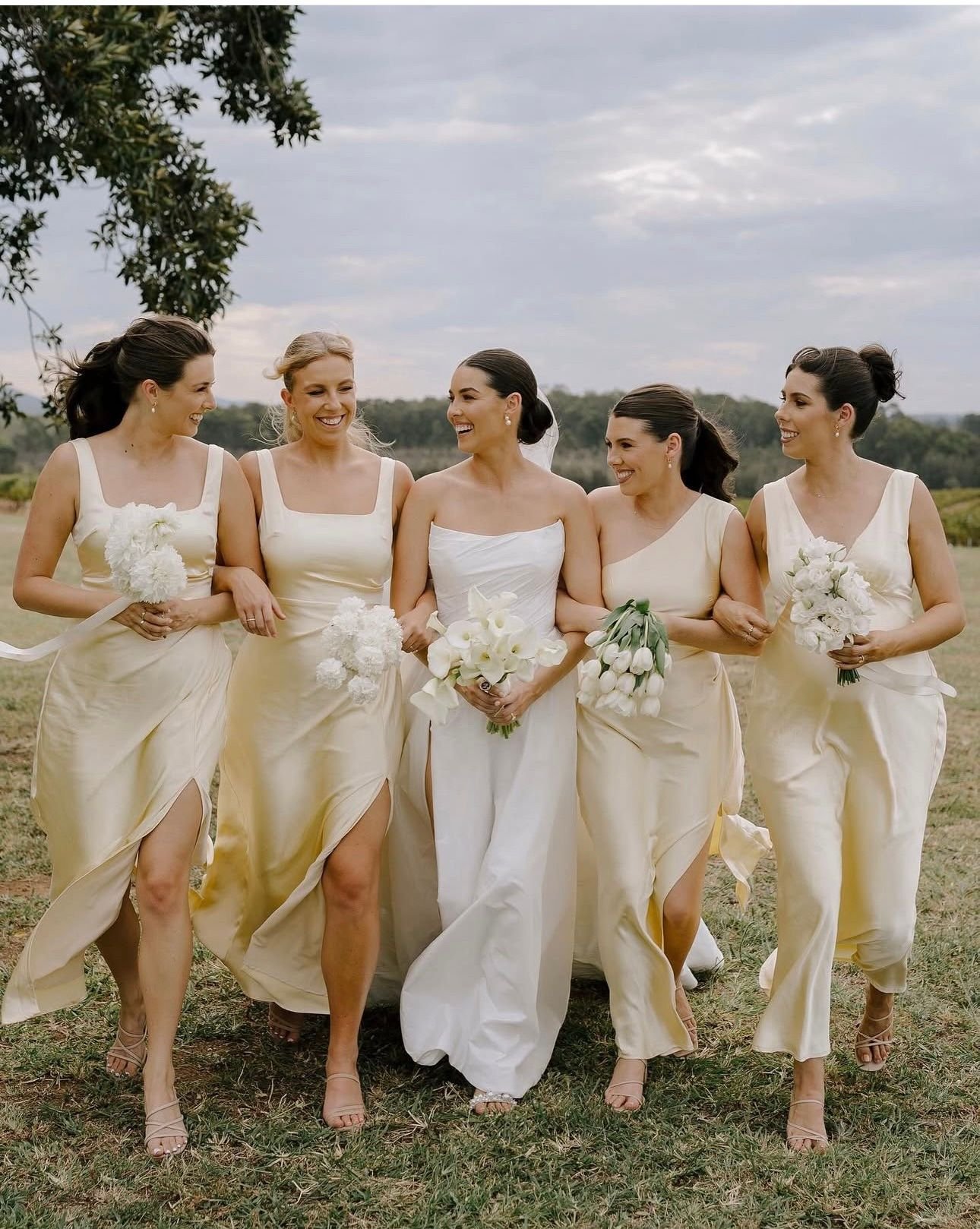A bride in a white gown and four bridesmaids in light yellow dresses walking outdoors, carrying white bouquets, smiling and interacting with each other, with greenery in the background.
