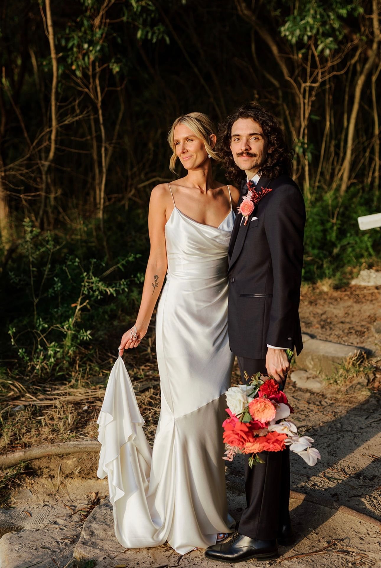 Bride and groom posing outdoors, bride in a white dress holding a bouquet, groom in a dark suit with floral boutonniere.