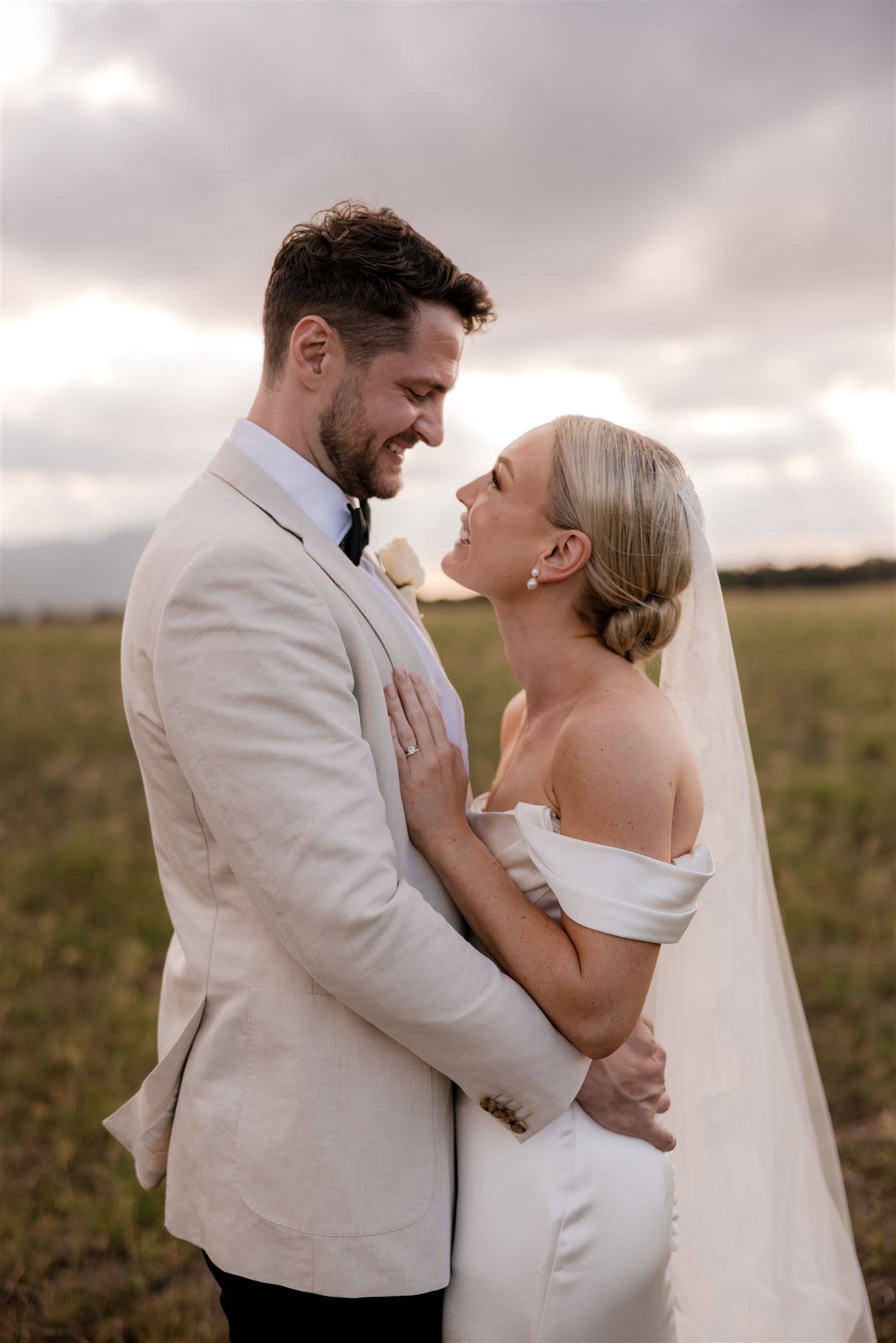 Bride and groom embracing outdoors in a field, wearing wedding attire, smiling at each other.
