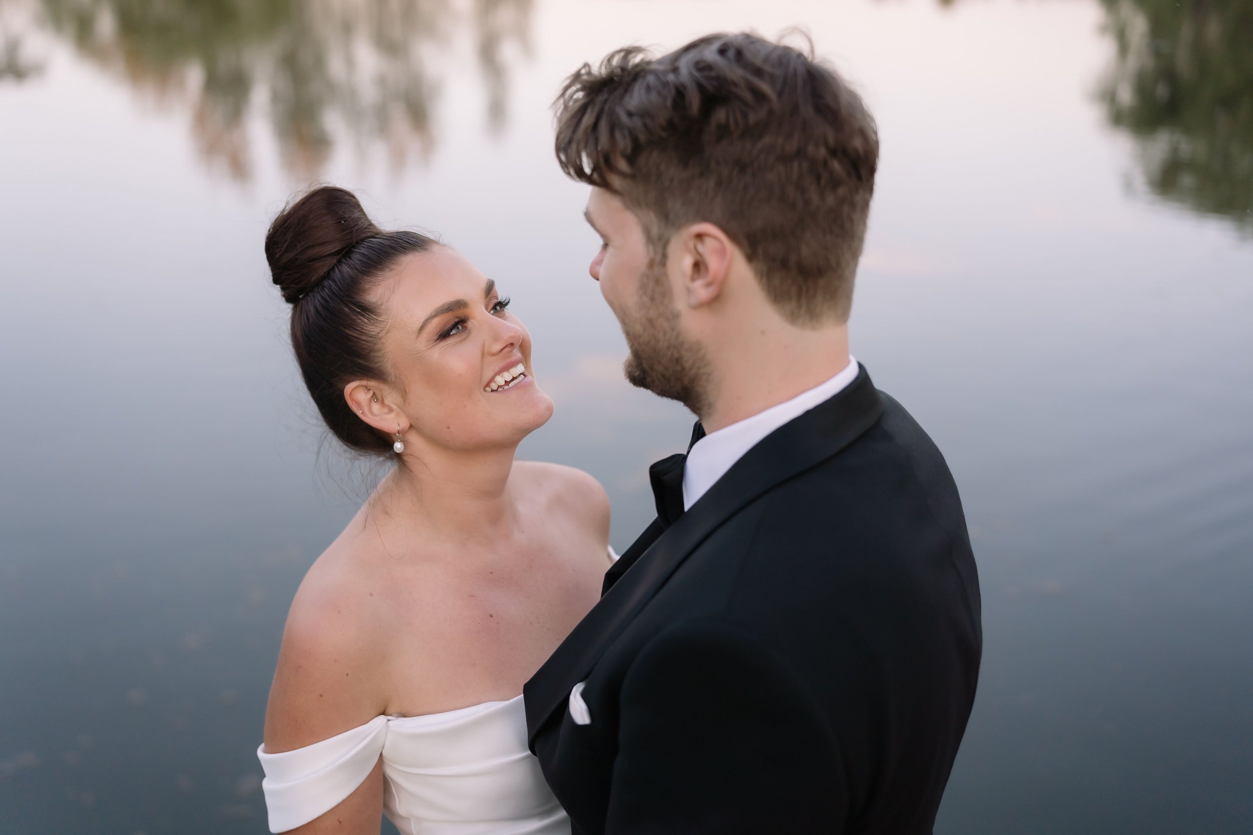 A couple in formal attire smiling at each other by a lake.