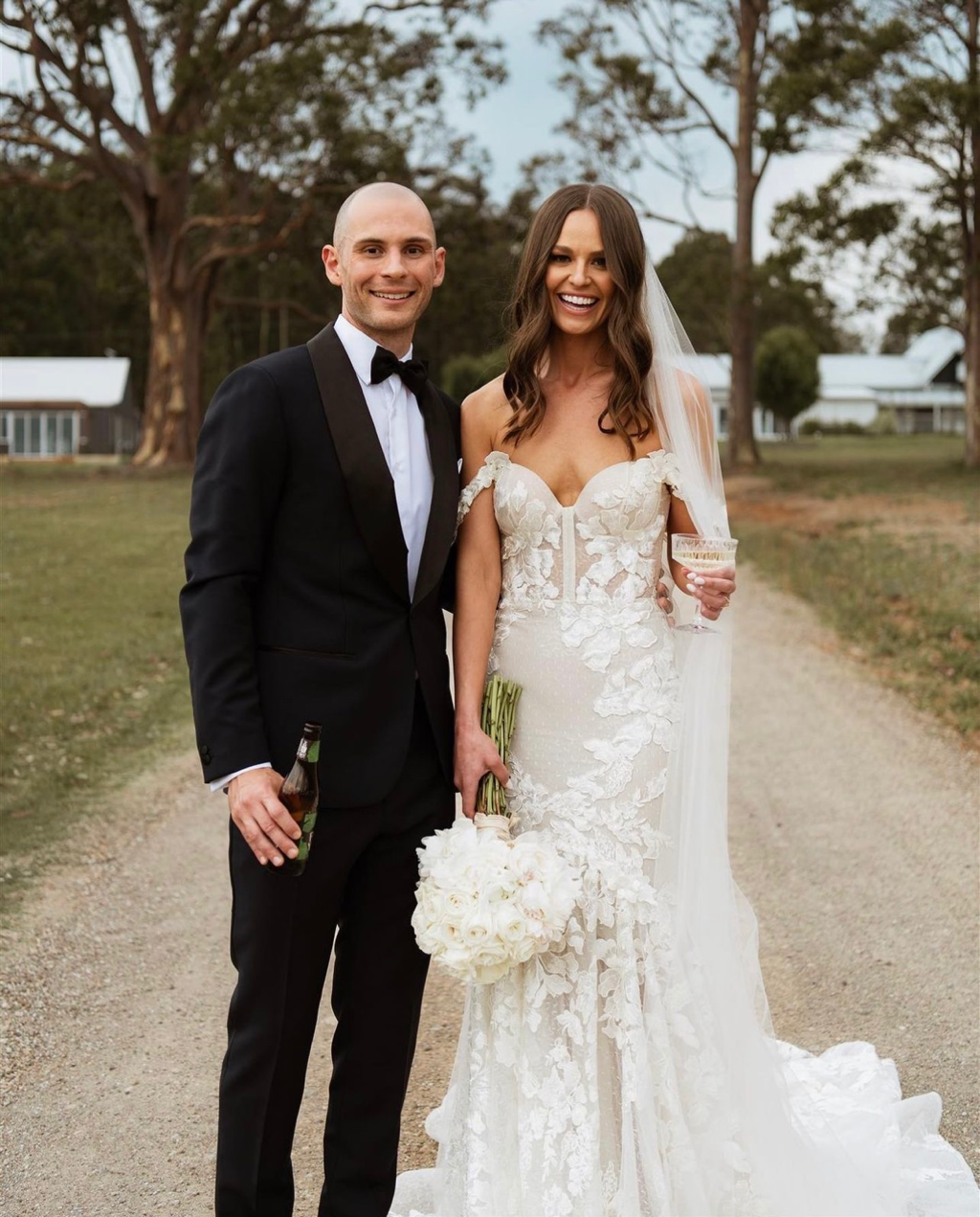Smiling bride and groom standing on a rural path, bride holds a bouquet and groom holds a drink, wearing wedding attire.
