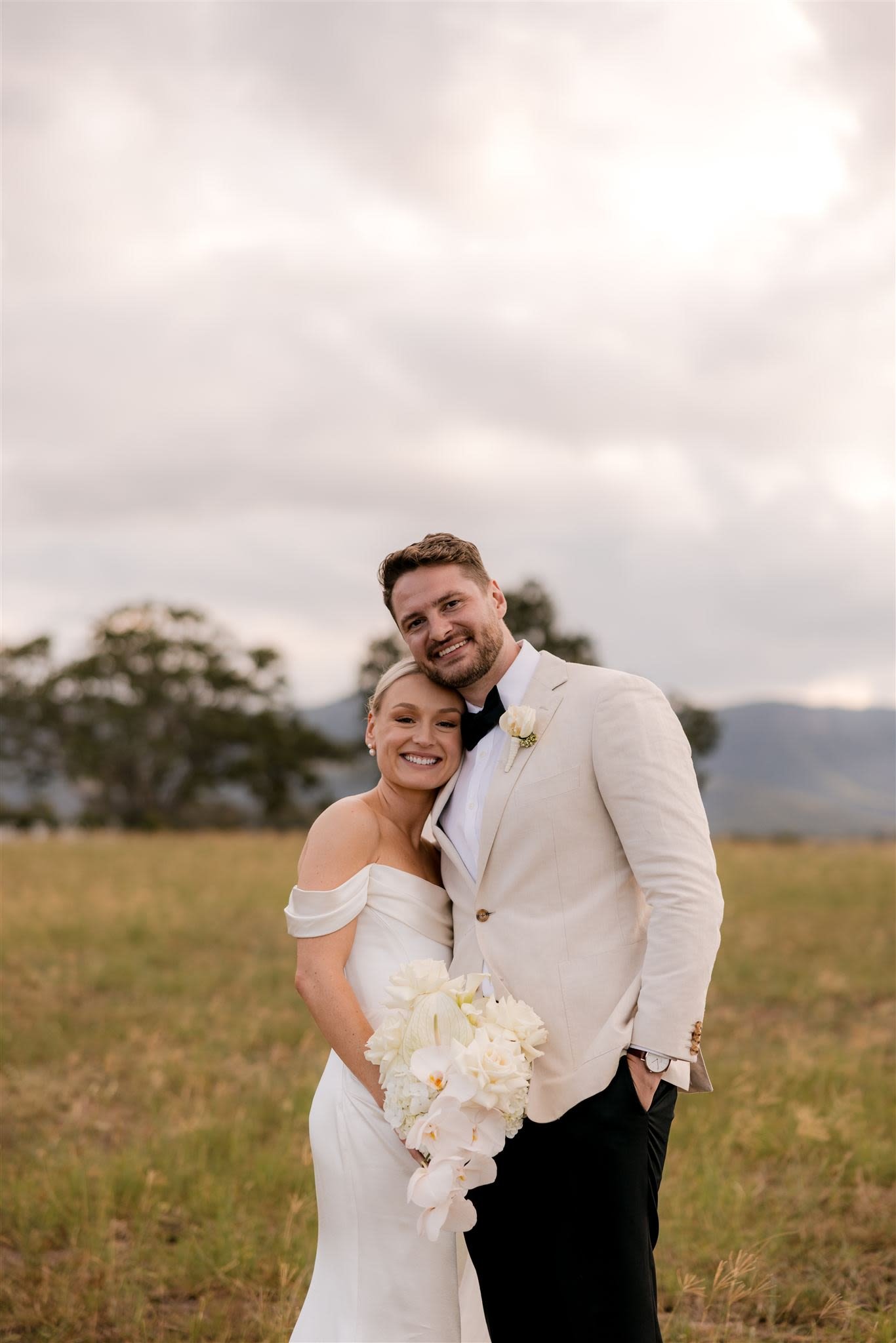 A bride and groom embracing outdoors, in a field with mountains in the background. The bride is wearing an off-shoulder wedding dress and holding a bouquet of white flowers, while the groom is in a light-colored suit with a boutonniere.
