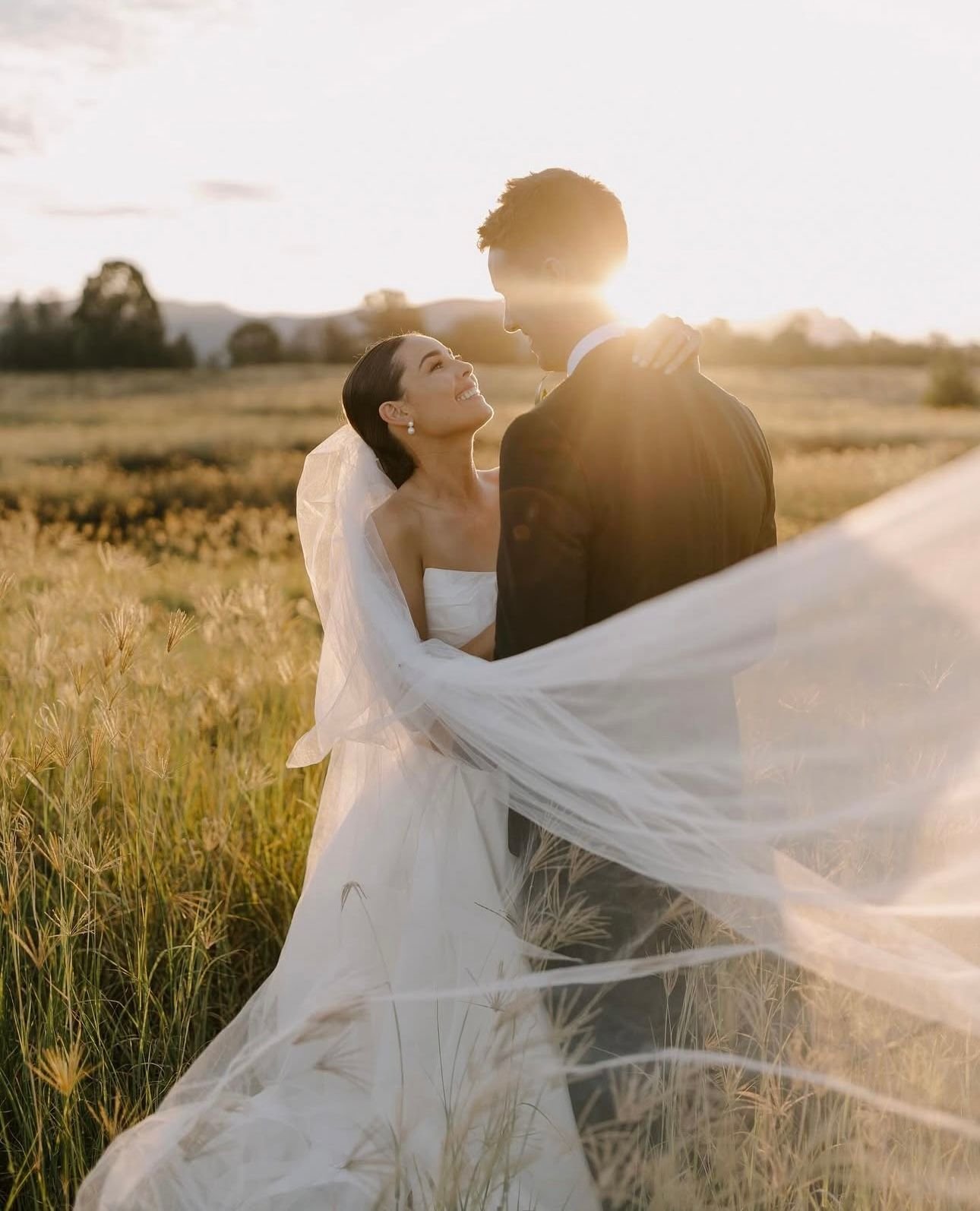 Bride and groom embracing in a sunlit field, bride wearing a long veil flowing in the breeze.