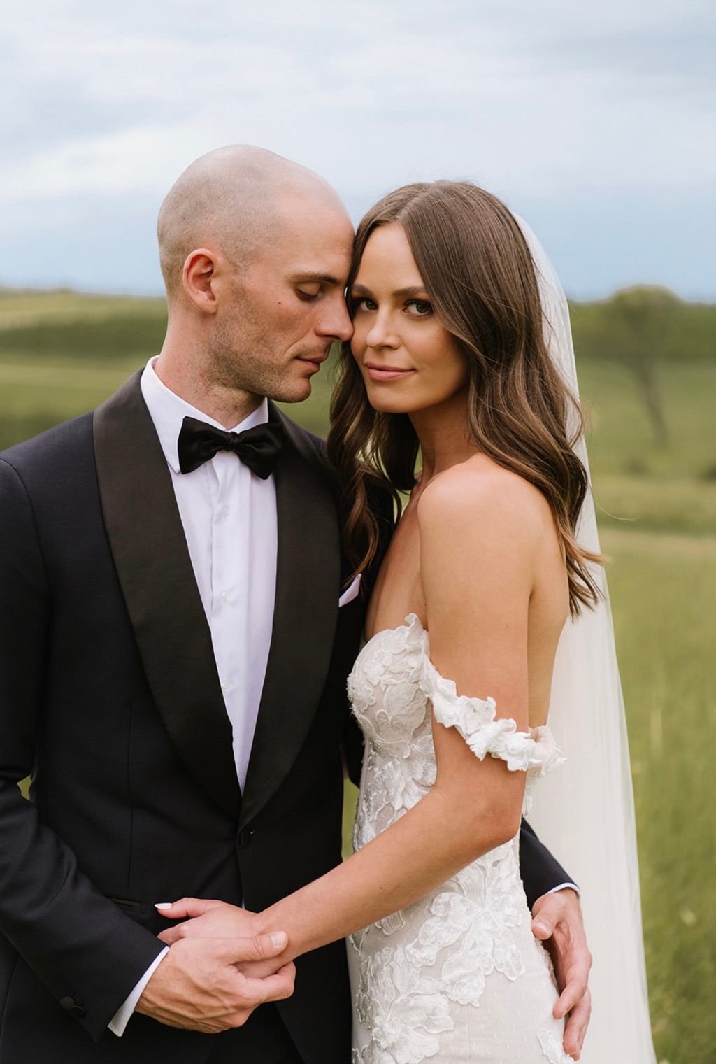 Bride and groom in wedding attire embracing outdoors with green field background.