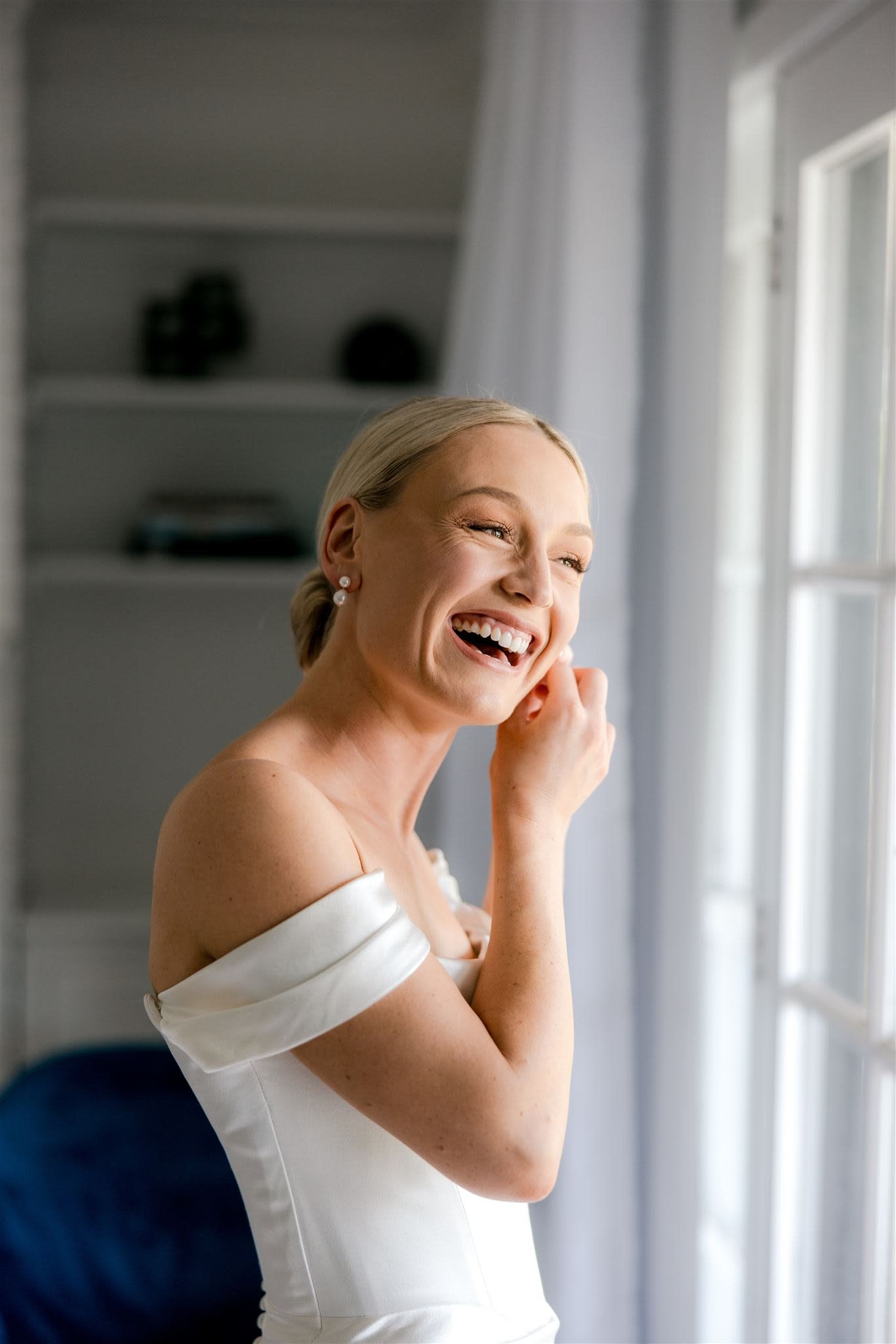 Bride smiling while wearing a white off-shoulder dress, adjusting her earring next to a window.