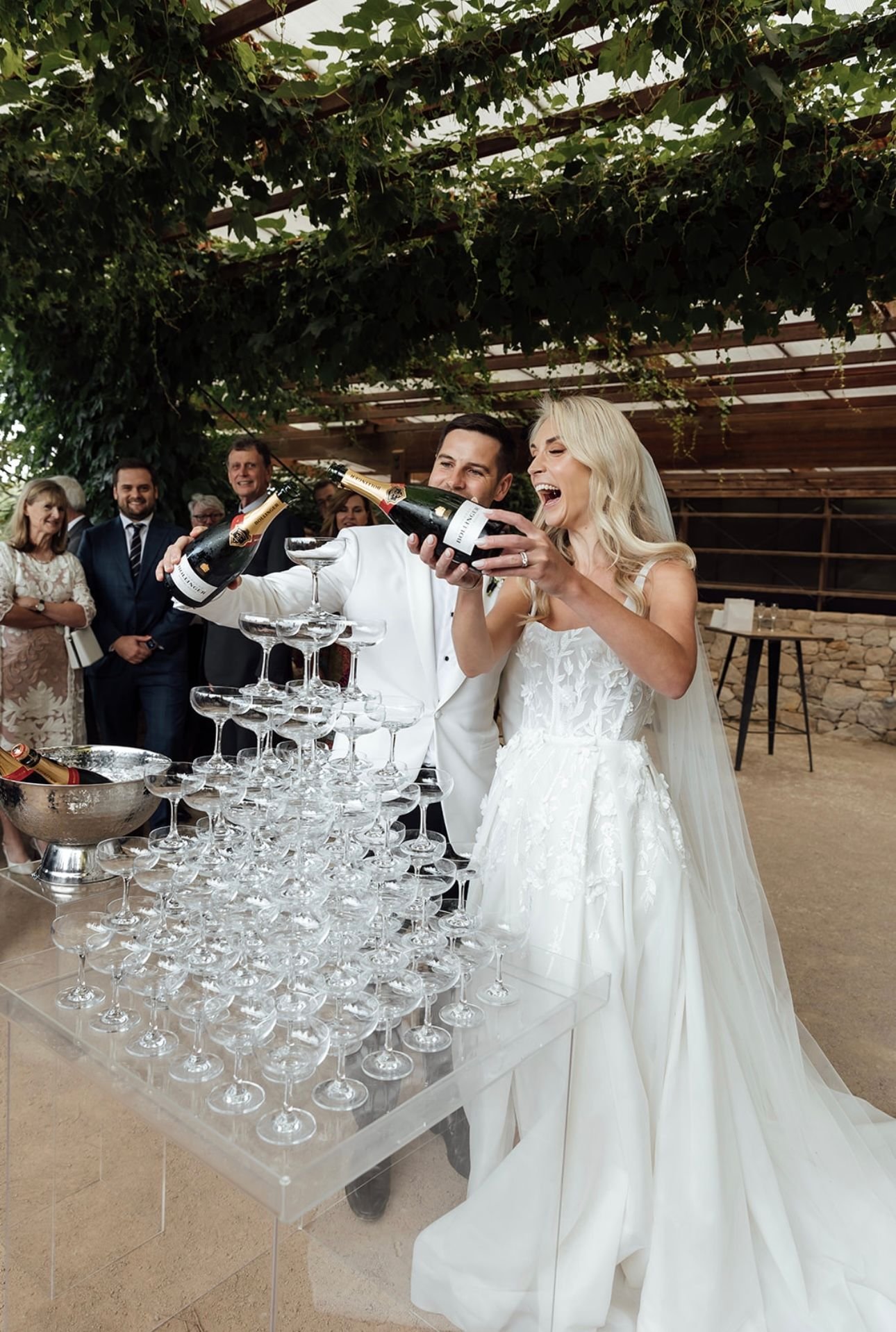 A bride and groom pouring champagne into a pyramid of glasses at a wedding reception, with guests watching under a canopy of greenery.