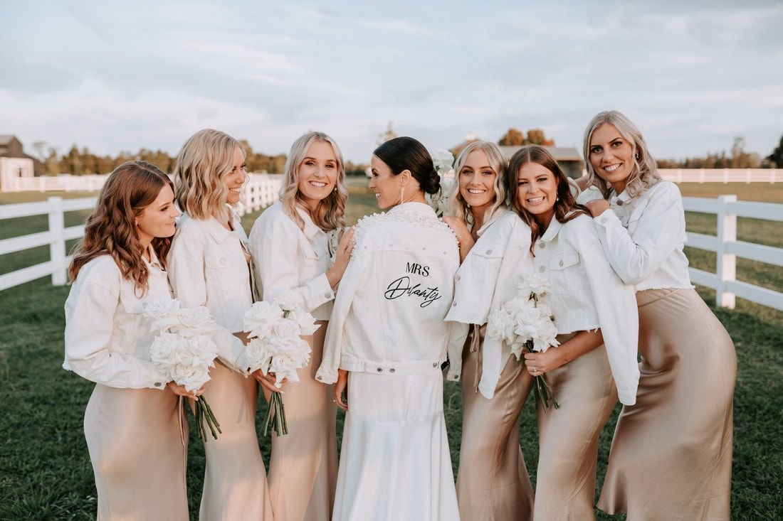 Bride with bridesmaids wearing white jackets and holding white flowers, posing outdoors near white fence.