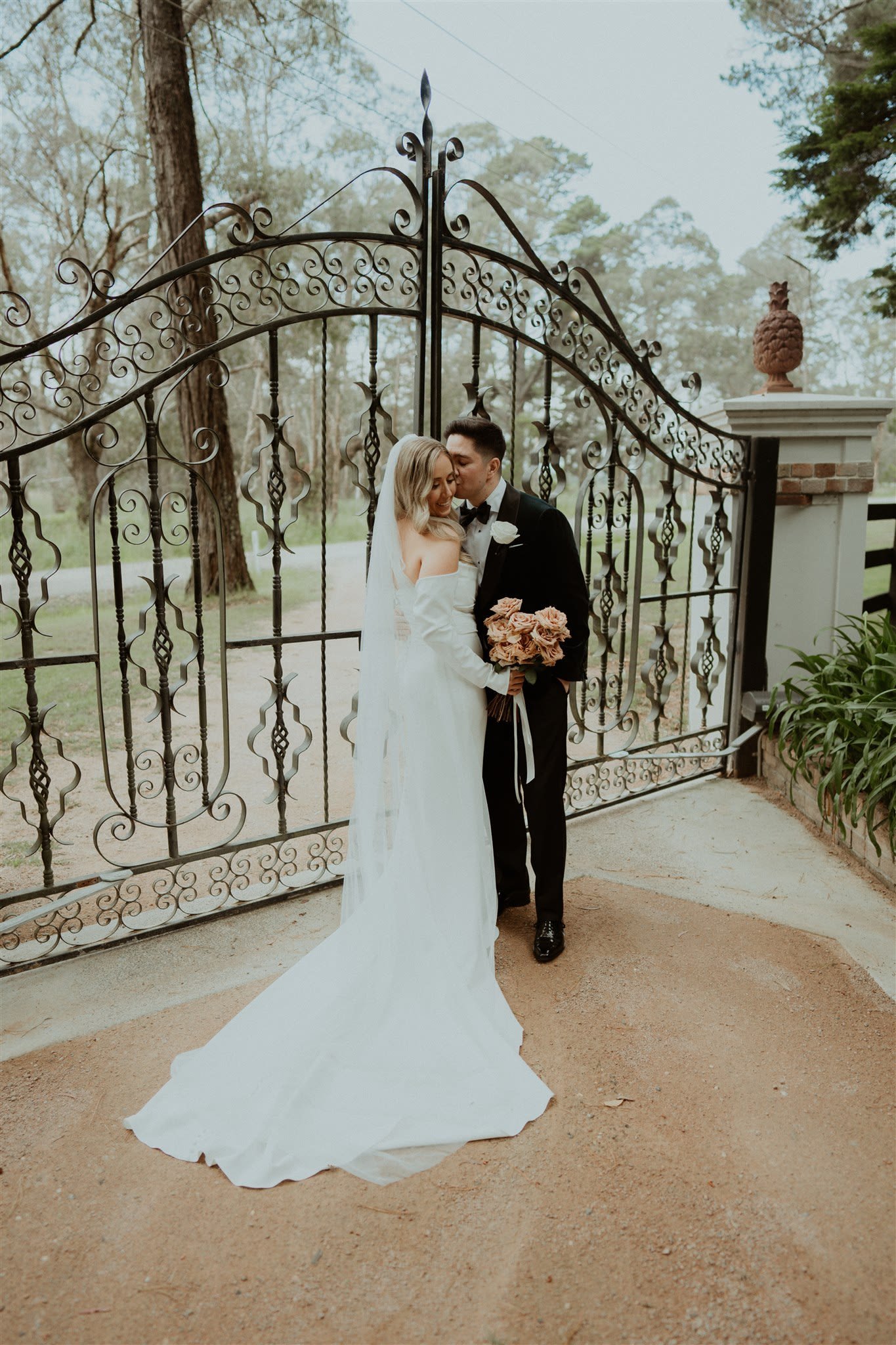 Bride and groom standing in front of an ornate wrought iron gate, surrounded by trees, with the bride holding a bouquet and the groom wearing a tuxedo.