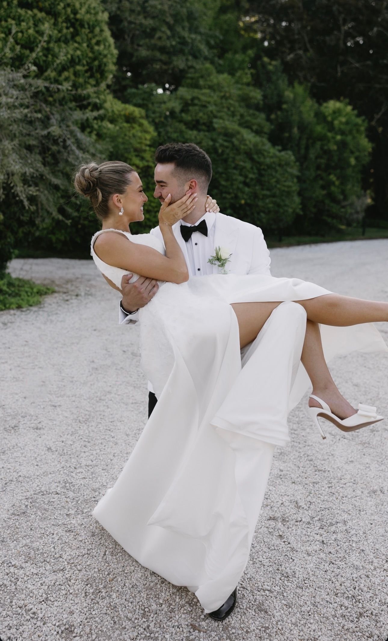 A groom in a white tuxedo holding a smiling bride wearing a white dress and heels on a gravel path with greenery in the background.