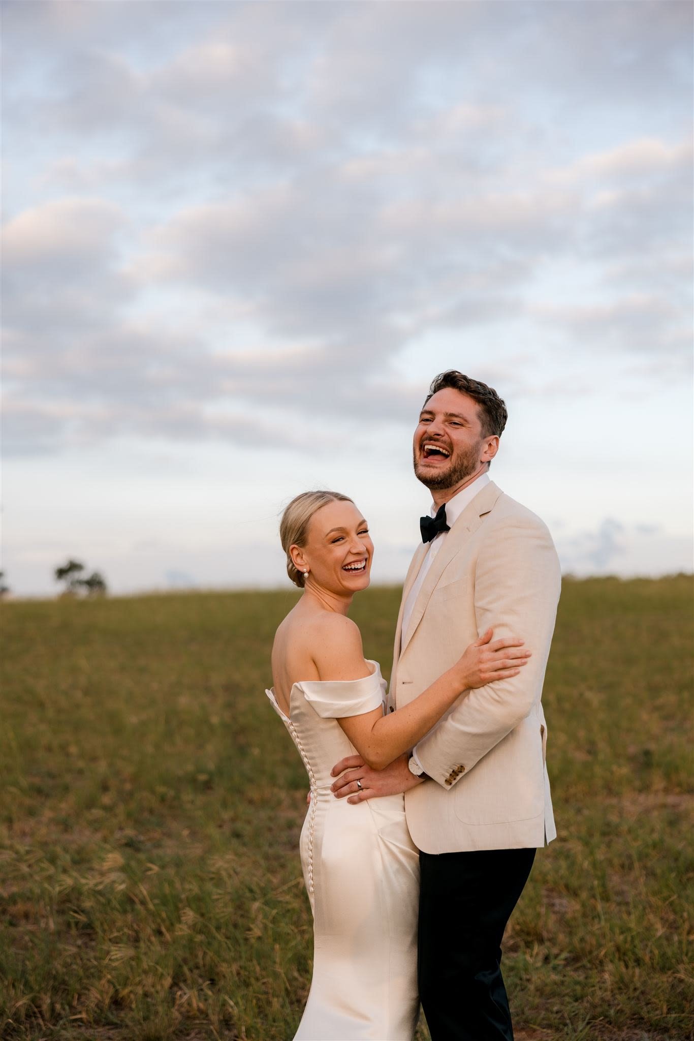 A joyful couple in wedding attire laughing and embracing in a grassy field under a cloudy sky.