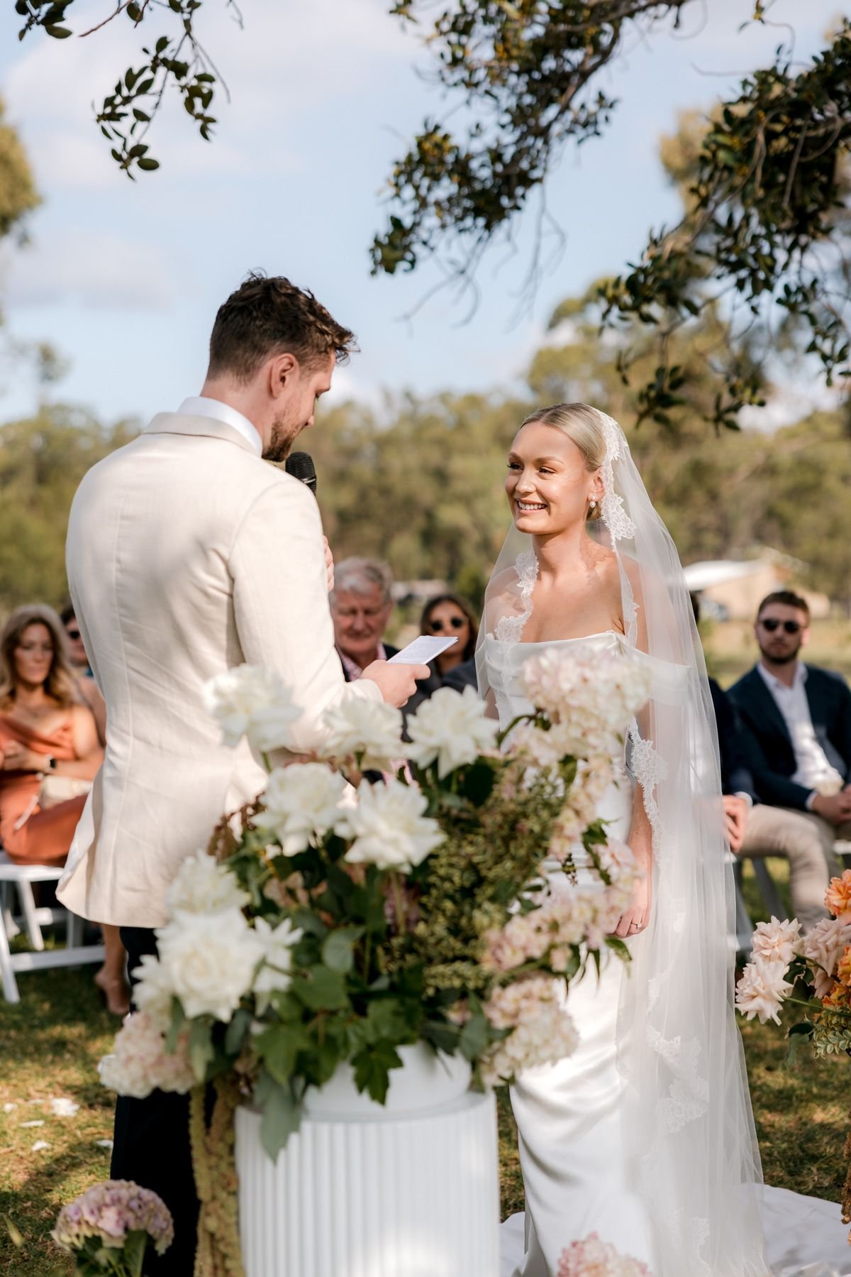 Outdoor wedding ceremony with a bride and groom exchanging vows, surrounded by guests and floral arrangements.