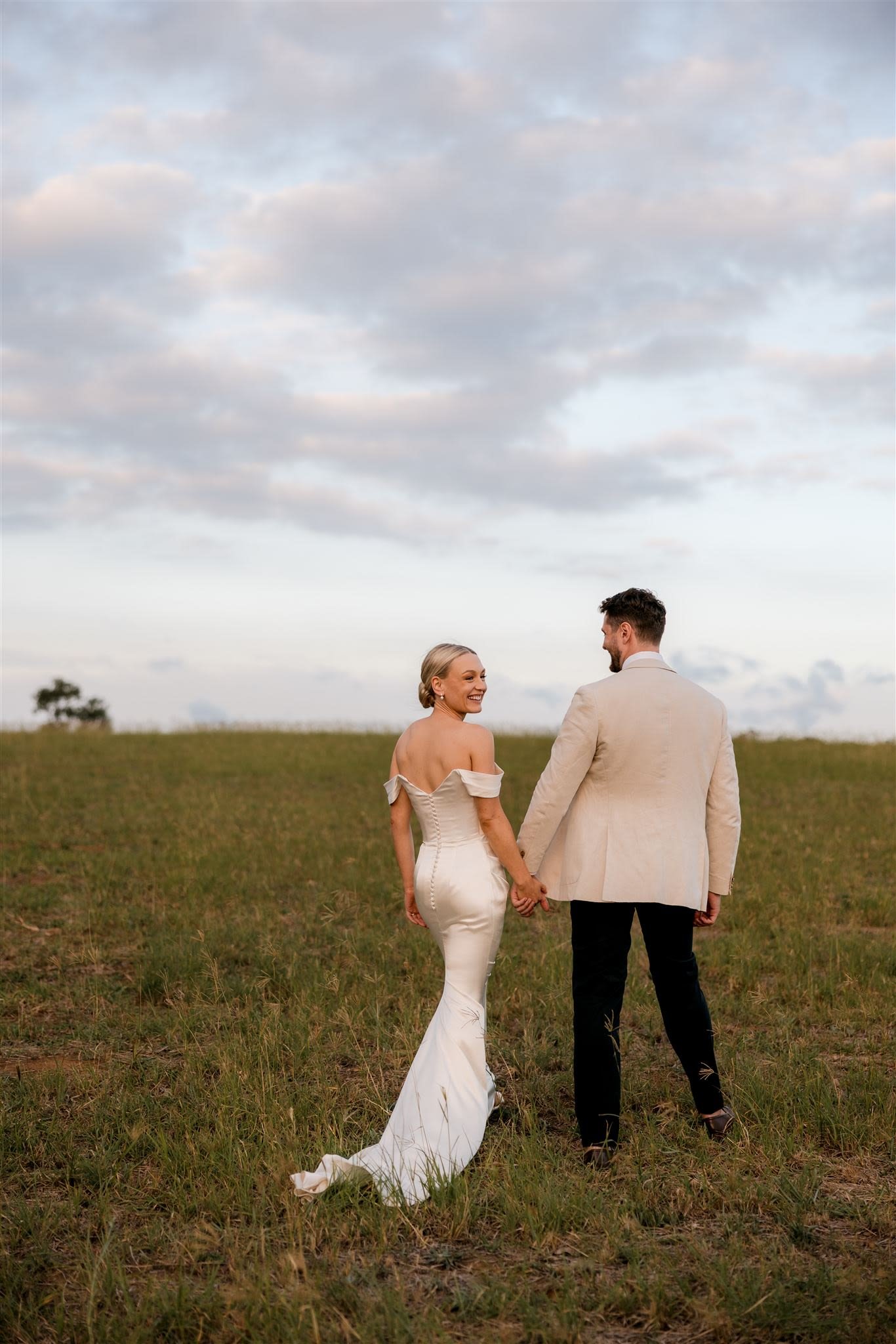 Couple holding hands, wearing formal attire, standing in a grassy field, cloudy sky overhead.