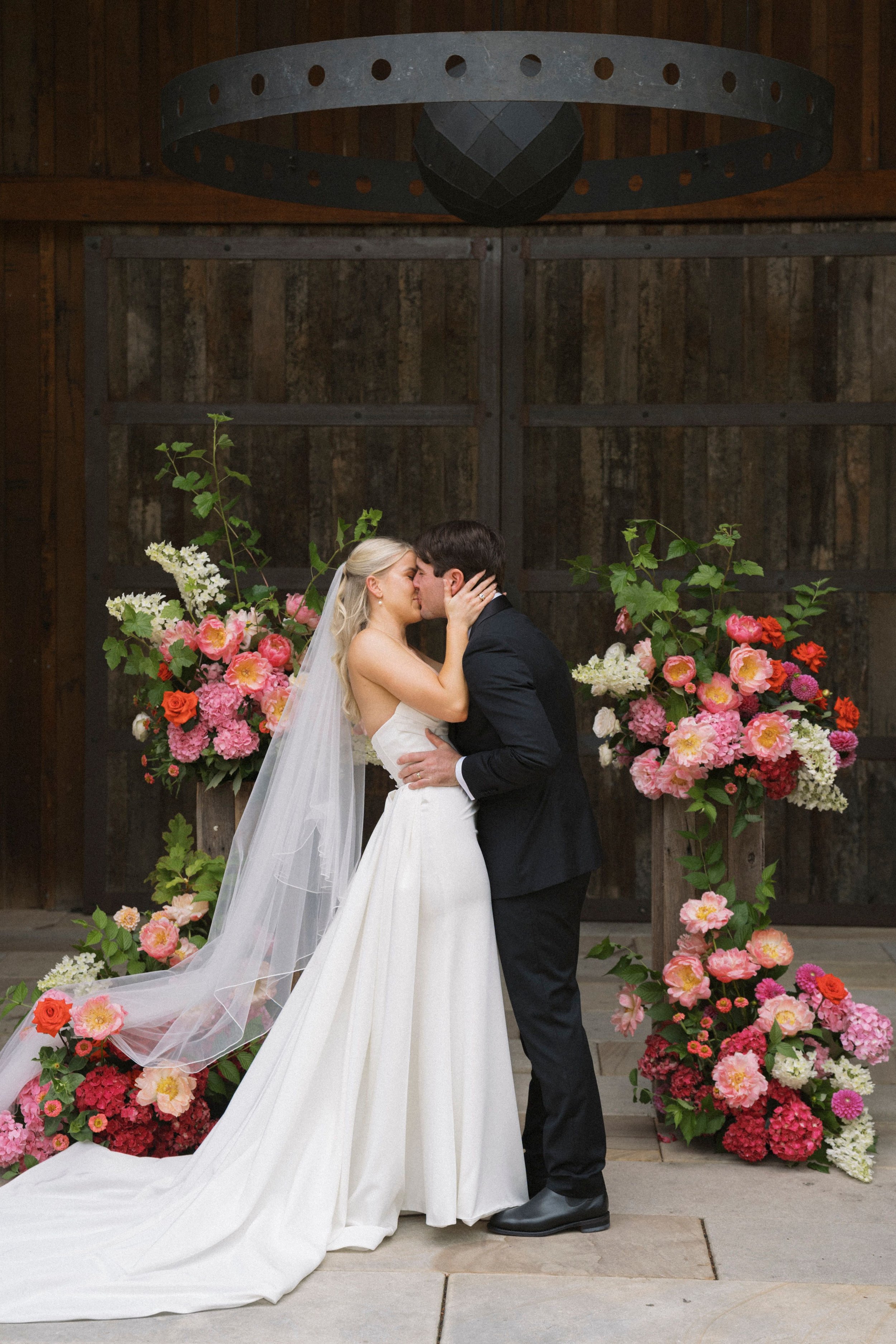 Bride and groom sharing a kiss at their wedding ceremony, surrounded by colorful floral arrangements, indoors with a rustic wooden backdrop.