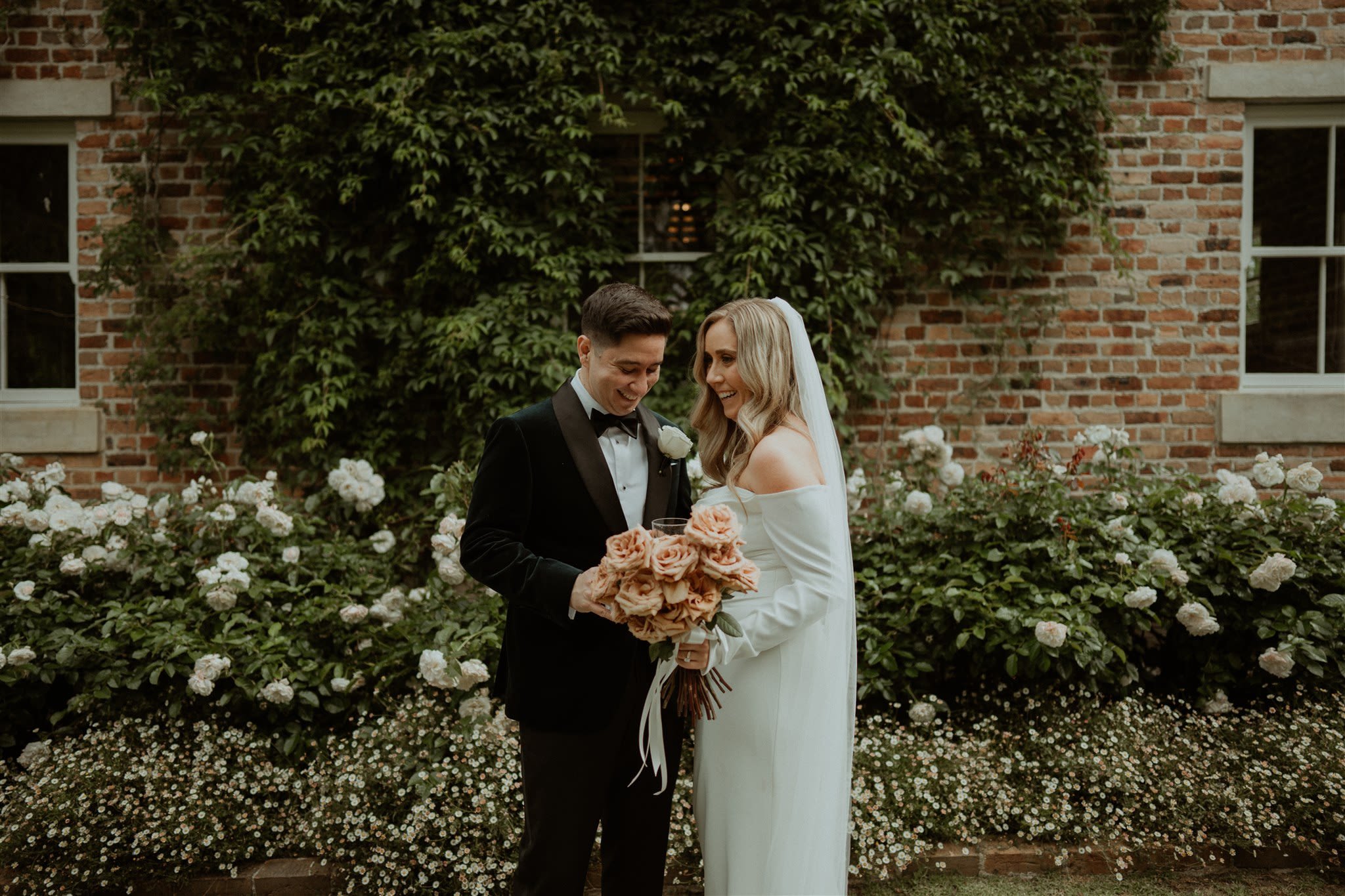 A bride and groom standing together in a garden in front of a brick wall, with the bride holding a bouquet of roses and wearing an off-the-shoulder white dress, and the groom in a black tuxedo with a white boutonniere.