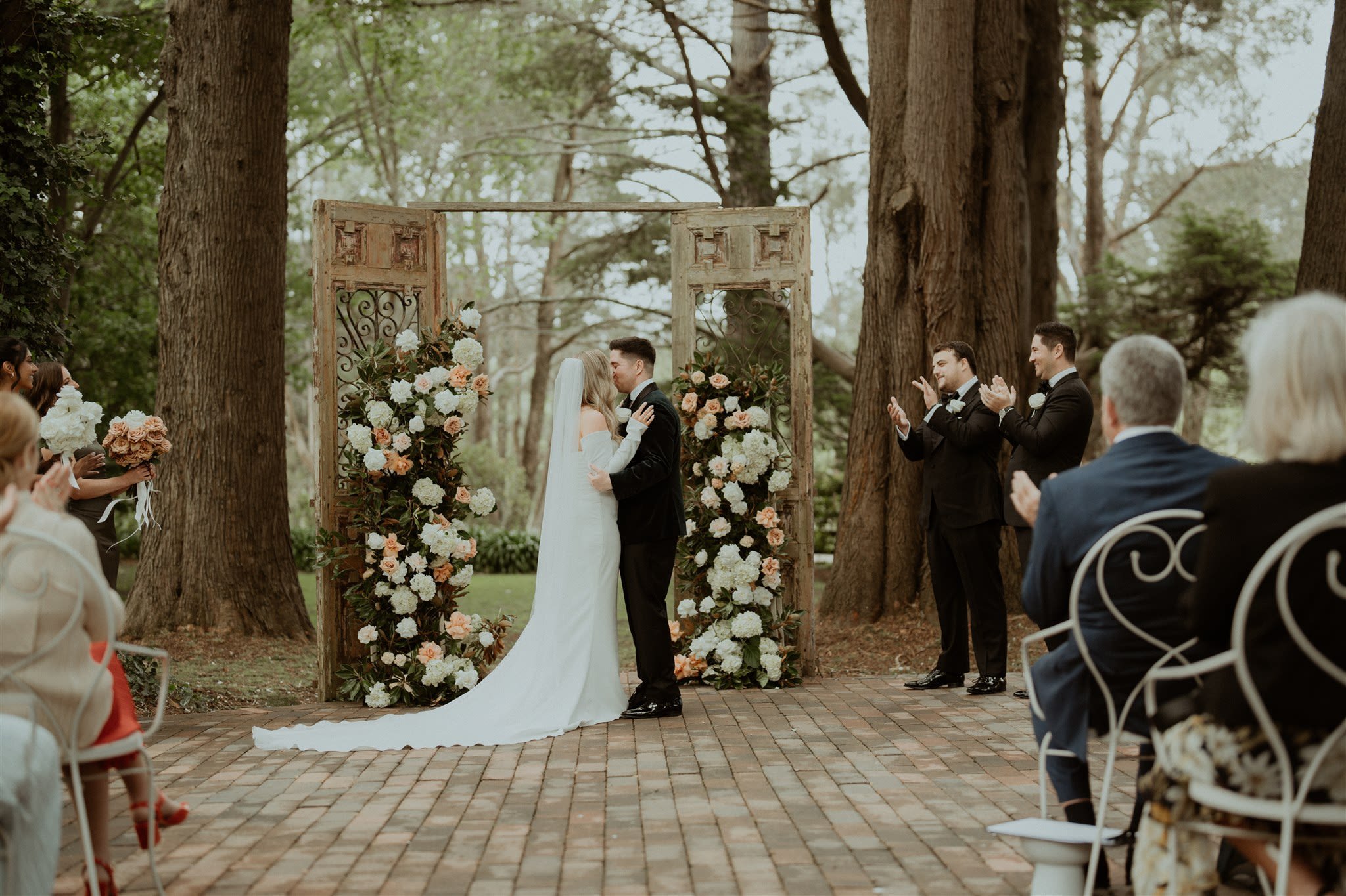 Bride and groom kissing at an outdoor wedding ceremony with floral decorations, surrounded by clapping guests and trees.