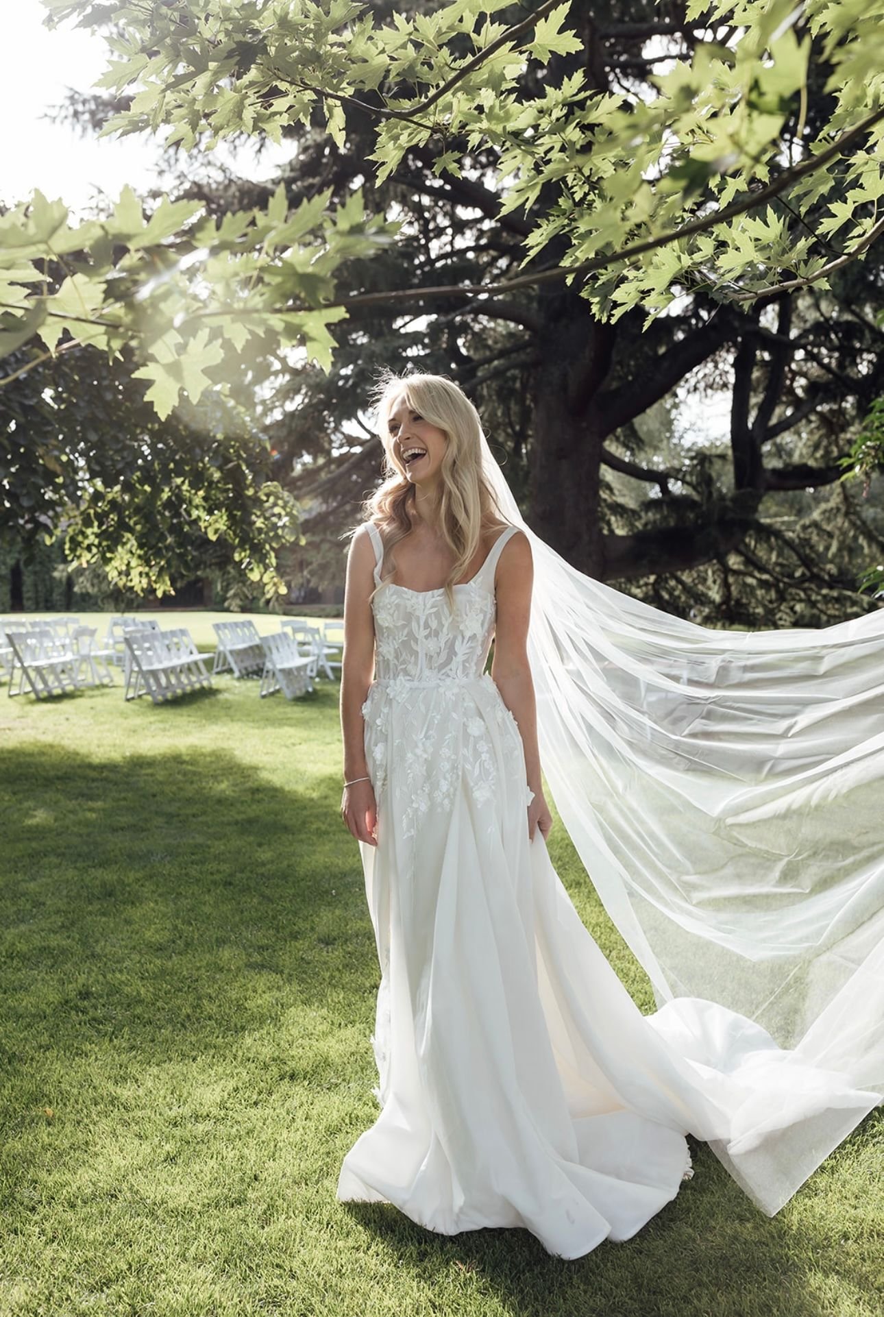 Bride in white wedding dress with a long veil, standing outdoors on grass, laughing under tree branches with sunlight filtering through.