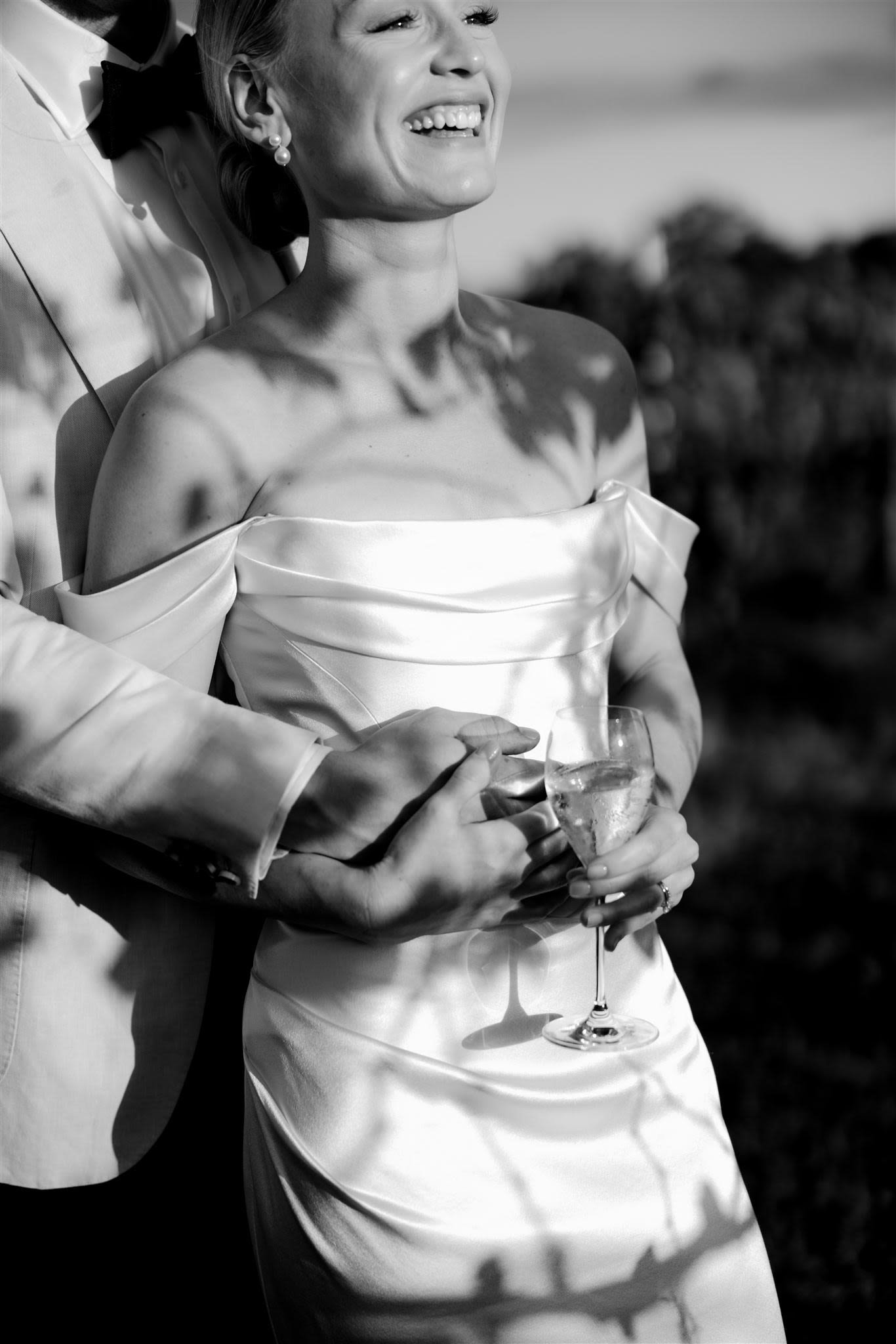 Bride smiling in a wedding dress, holding a wine glass, with shadow patterns on her dress and a person embracing her from behind.