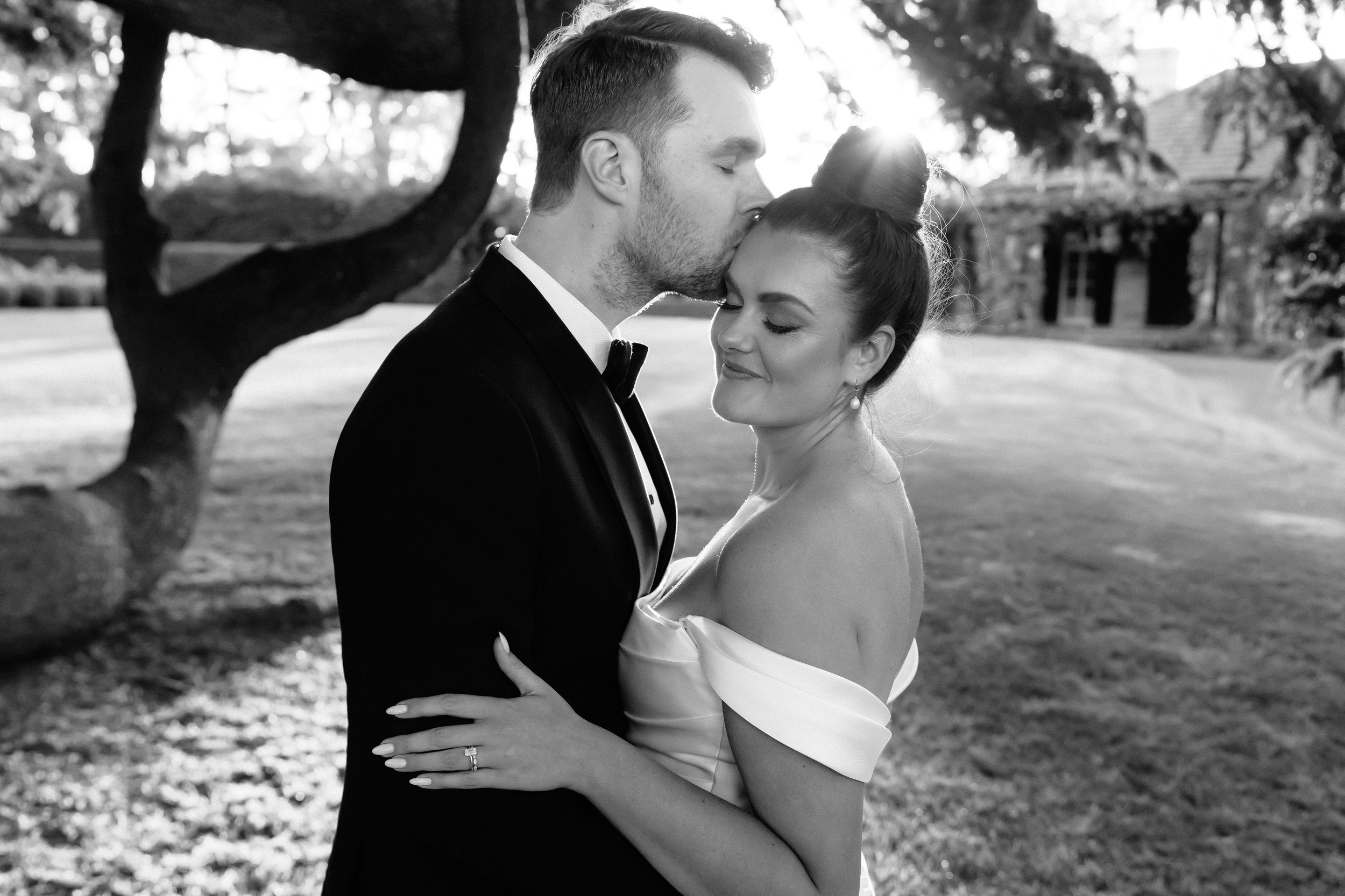 Black and white photo of a groom kissing a bride on the forehead in a garden setting, both in formal attire.