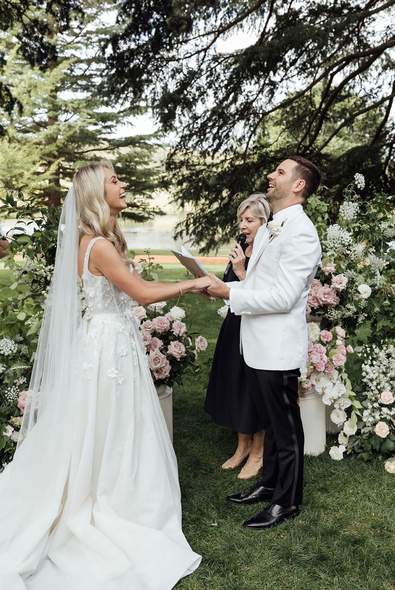 Bride and groom exchanging vows outdoors with officiant, surrounded by flowers and greenery.