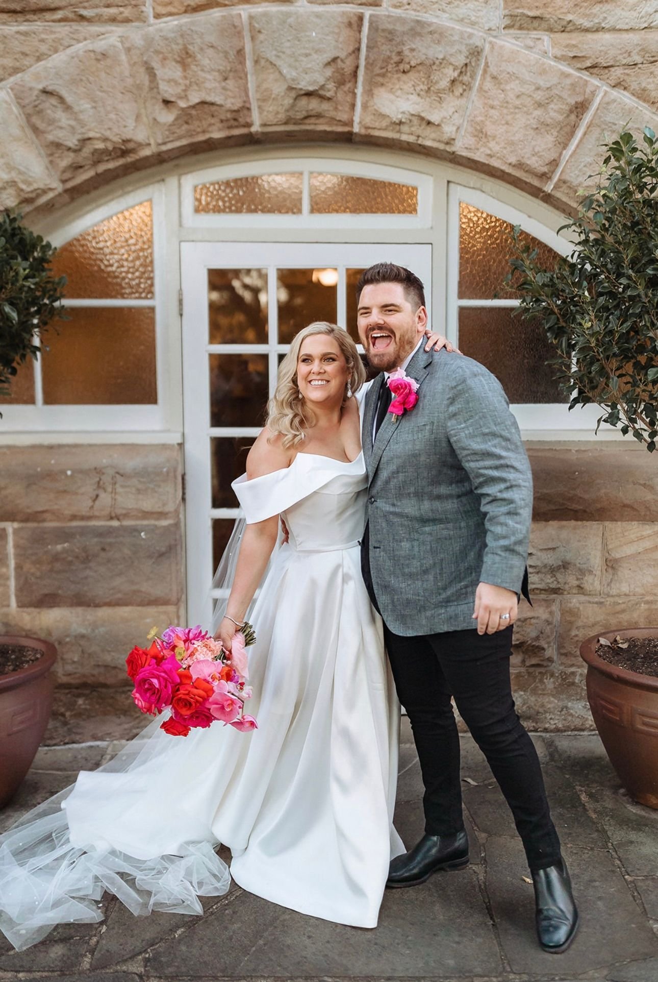 Bride and groom smiling in wedding attire in front of stone building.