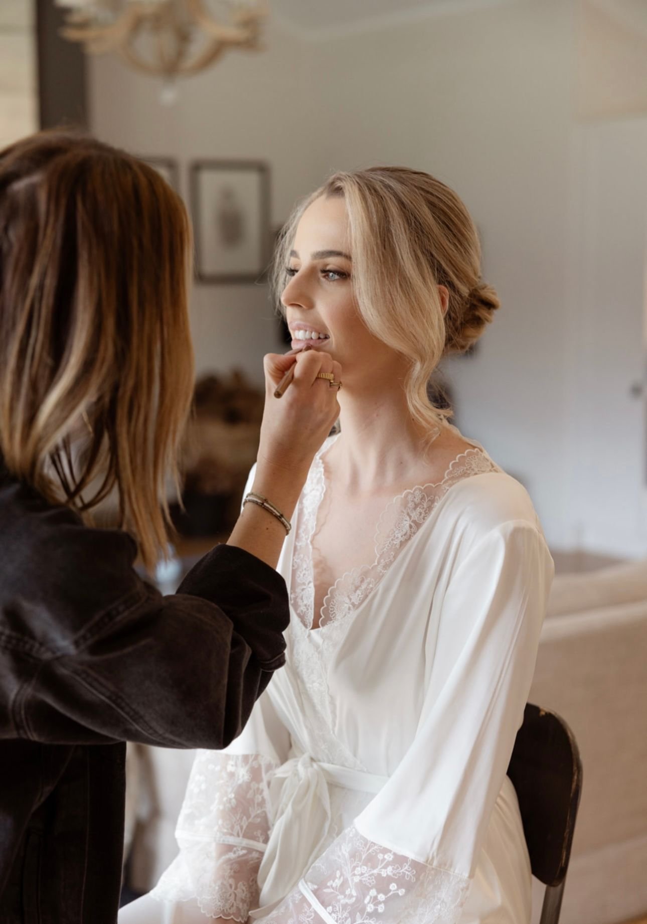 Bride in lace robe getting her makeup done by a stylist, preparing for a wedding.