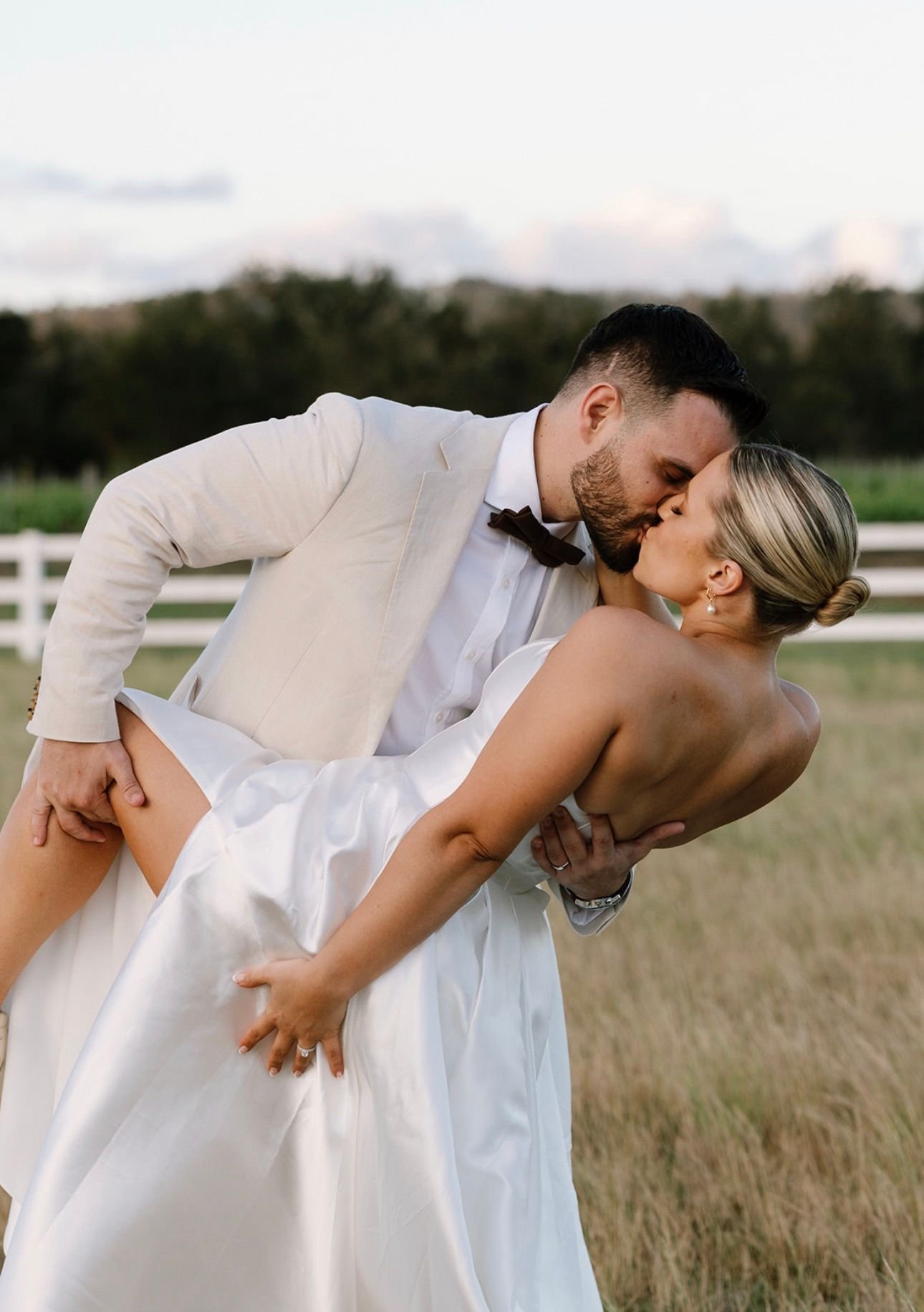 Bride and groom kissing, dressed in wedding attire, with groom holding bride in a dip pose outdoors.