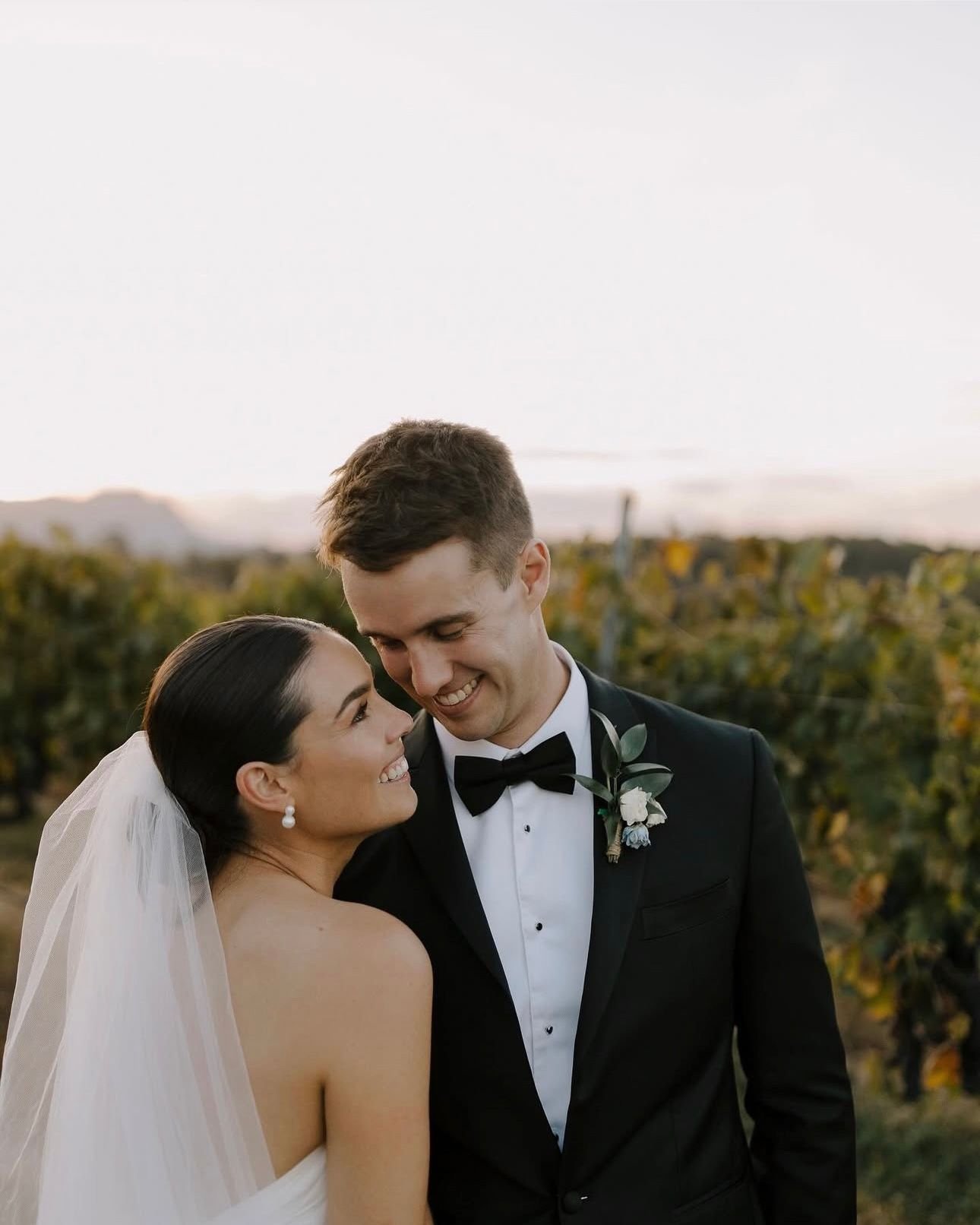 A bride and groom smiling at each other in an outdoor setting, wearing formal wedding attire. The bride is in a strapless dress with a veil, and the groom is in a black tuxedo with a boutonniere. The backdrop features a natural landscape.