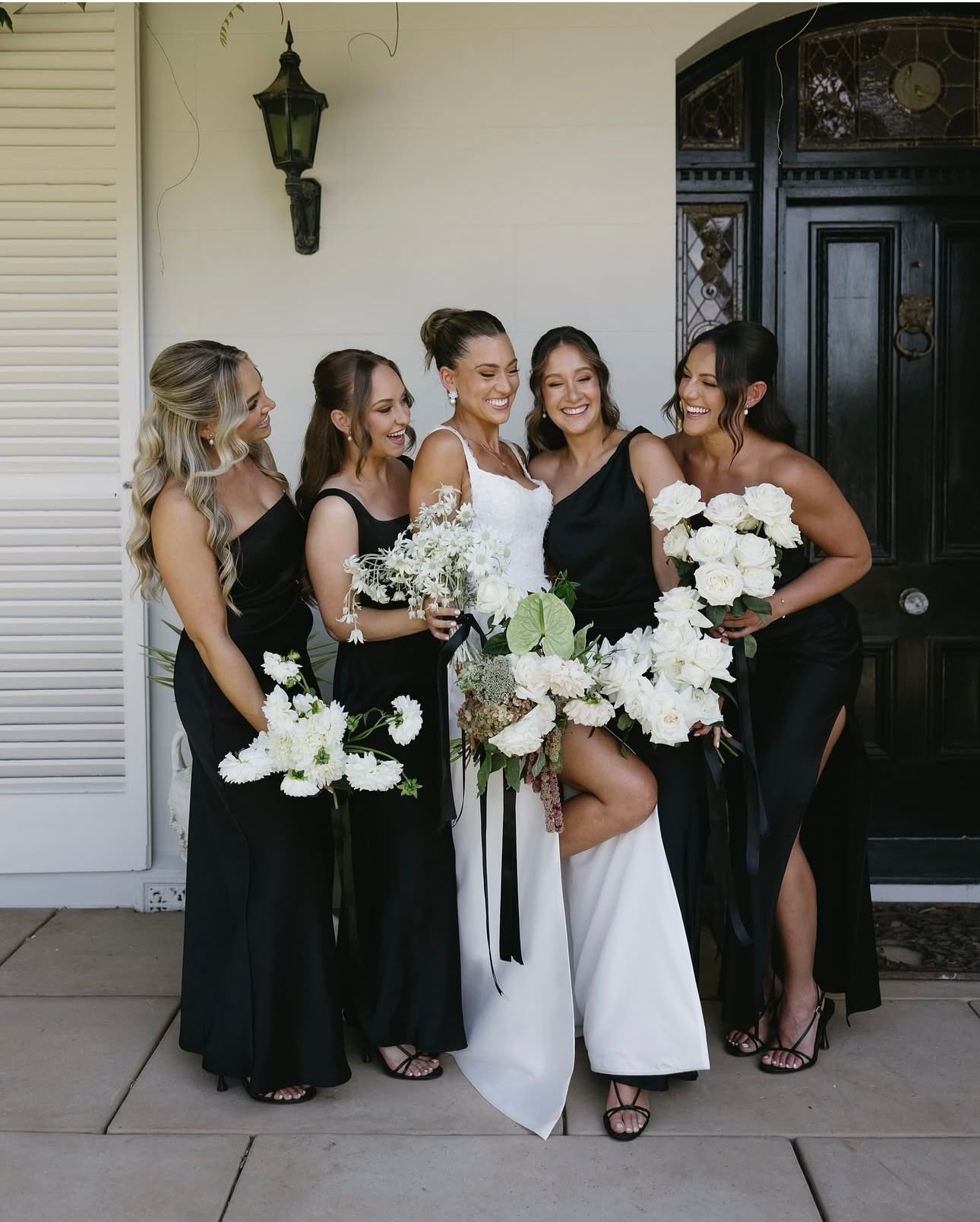A bride in a white gown posing with four bridesmaids in black dresses, all holding white floral bouquets, standing in front of a dark door.