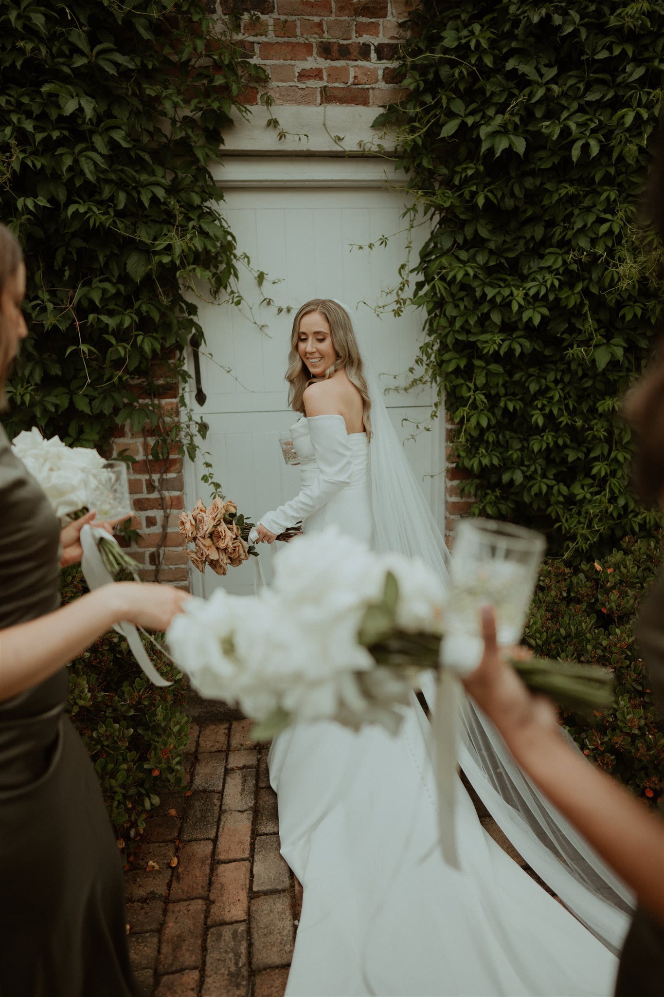 Bride in white dress holding bouquet, walking on brick path surrounded by greenery and bridesmaids with flowers.