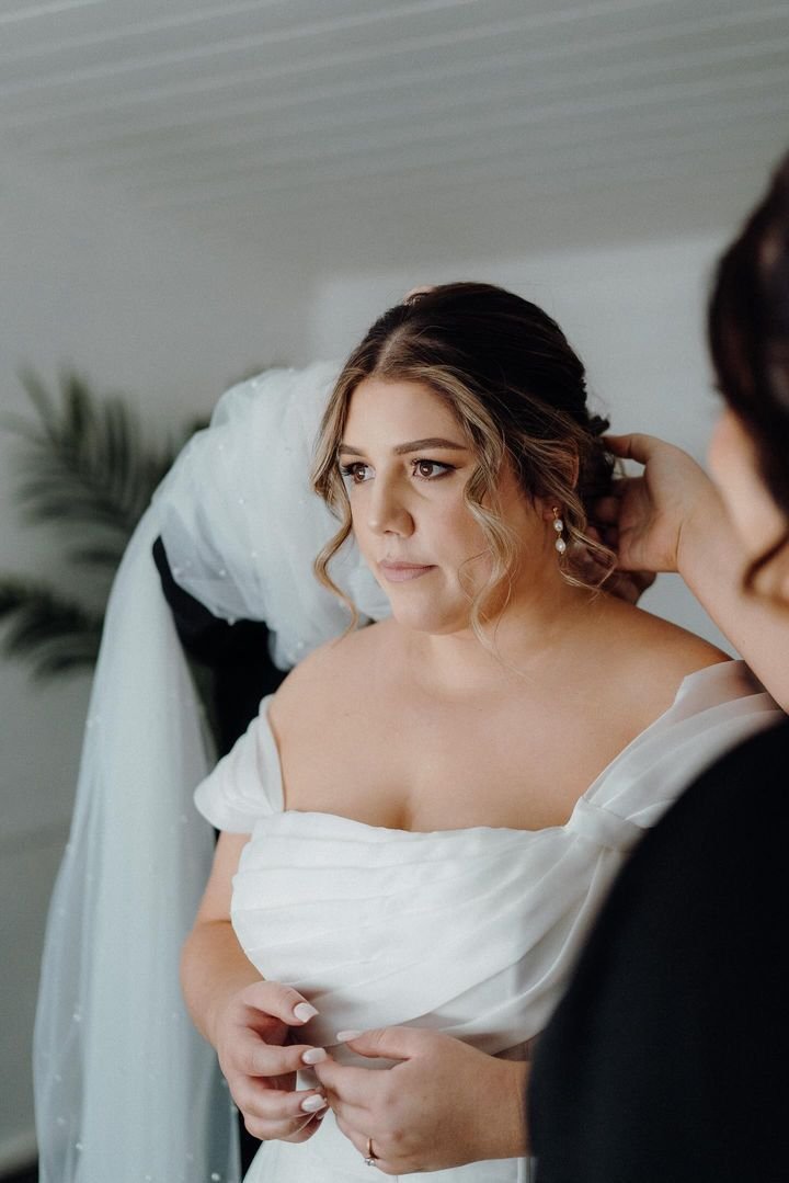 Bride wearing a white off-shoulder wedding dress being attended to by another person.