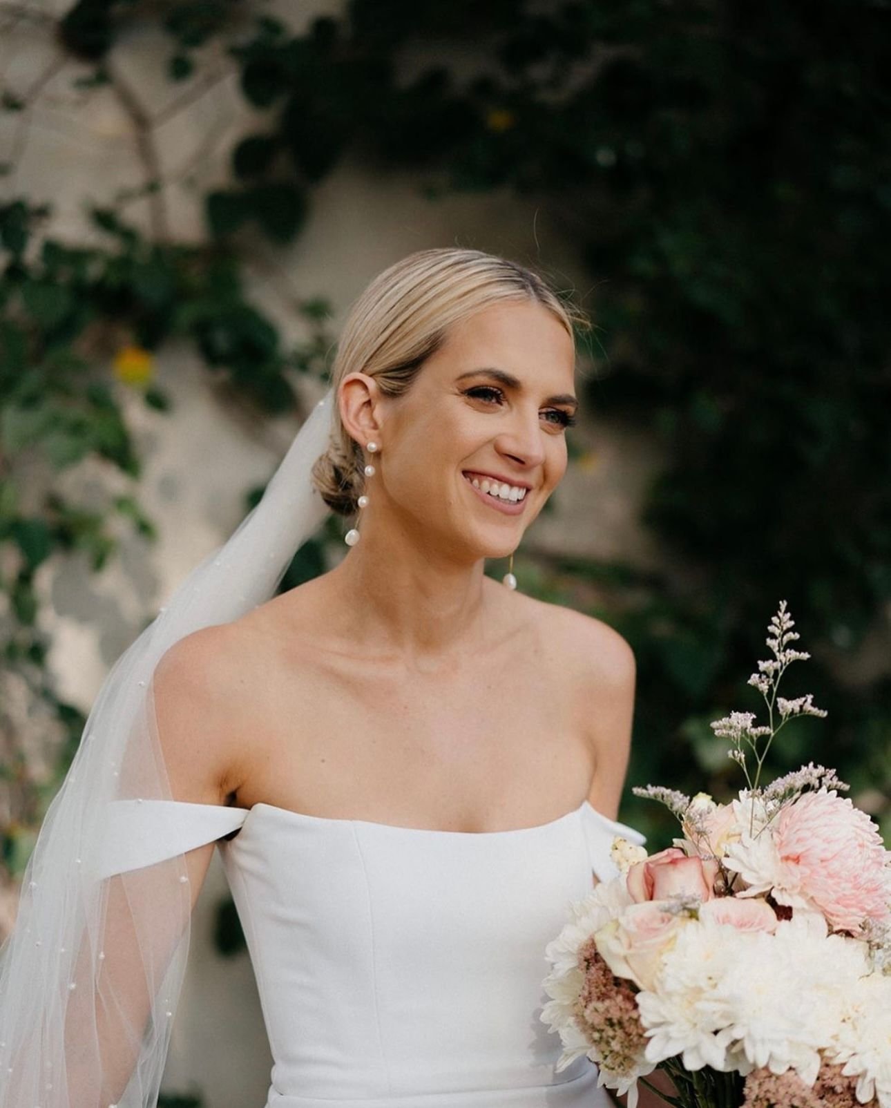 Bride in white wedding dress with veil and bouquet in outdoor setting.