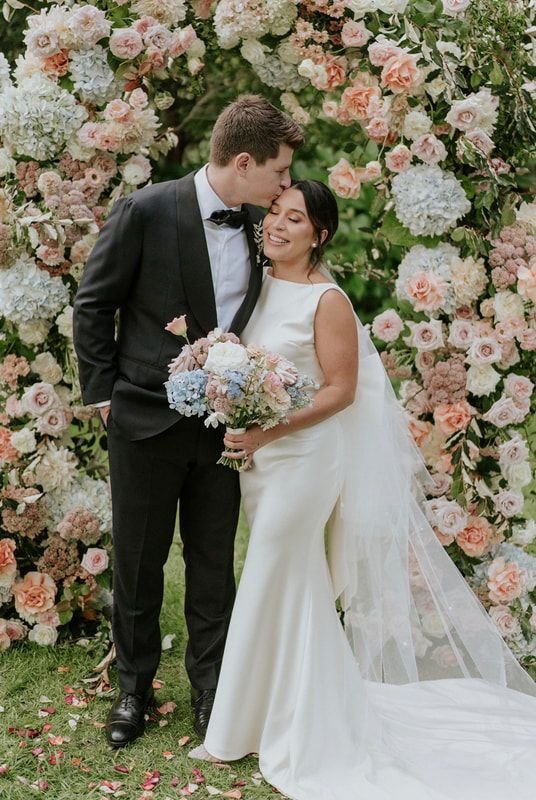 Bride and groom under a floral arch, bride in white gown holding bouquet, groom in black suit and bow tie, outdoor wedding setting.
