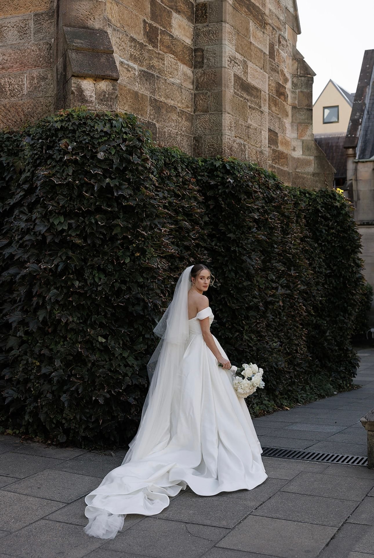 Bride in a white wedding dress and veil holding a bouquet, standing near an ivy-covered wall and stone building.