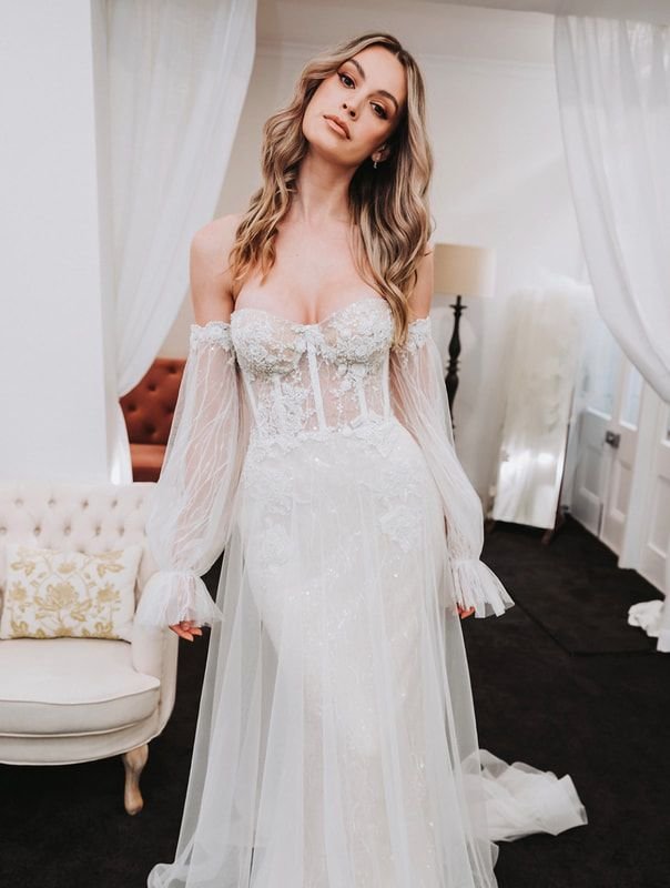Woman in bridal gown with sheer long sleeves, standing in a room with decor including a white armchair and curtains.