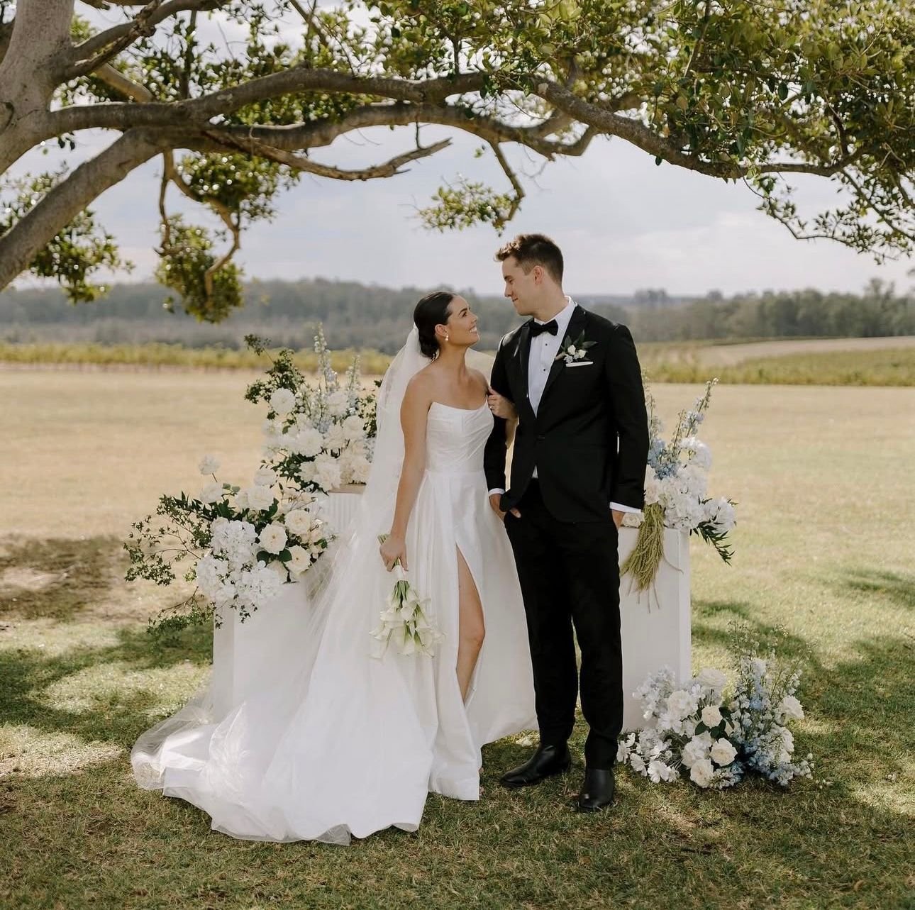 Bride and groom standing outdoors under a tree, with floral arrangements and a scenic landscape.