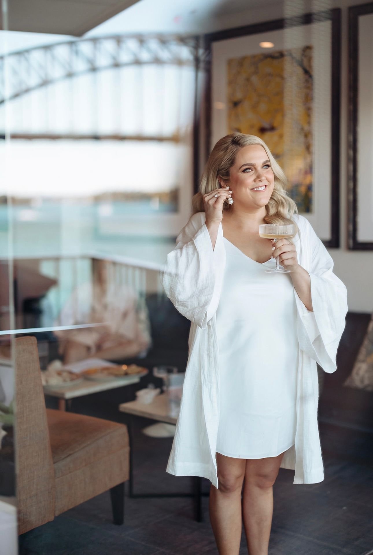 Woman in white dress holding a glass and wearing a robe, standing in a room with large windows.