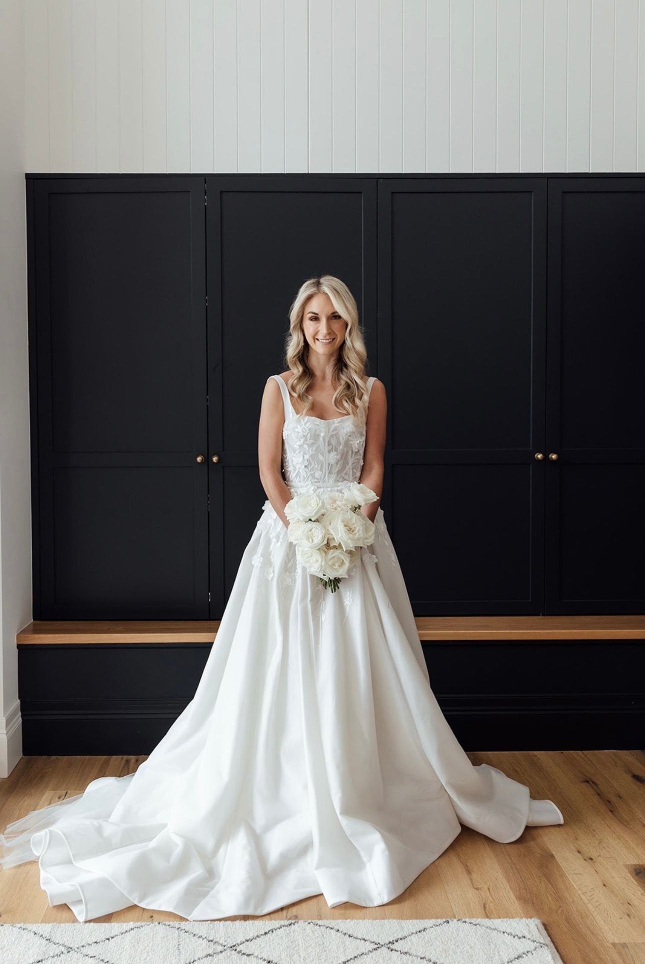 Bride in a white wedding dress holding a bouquet of white roses, standing indoors against a black cabinet background.